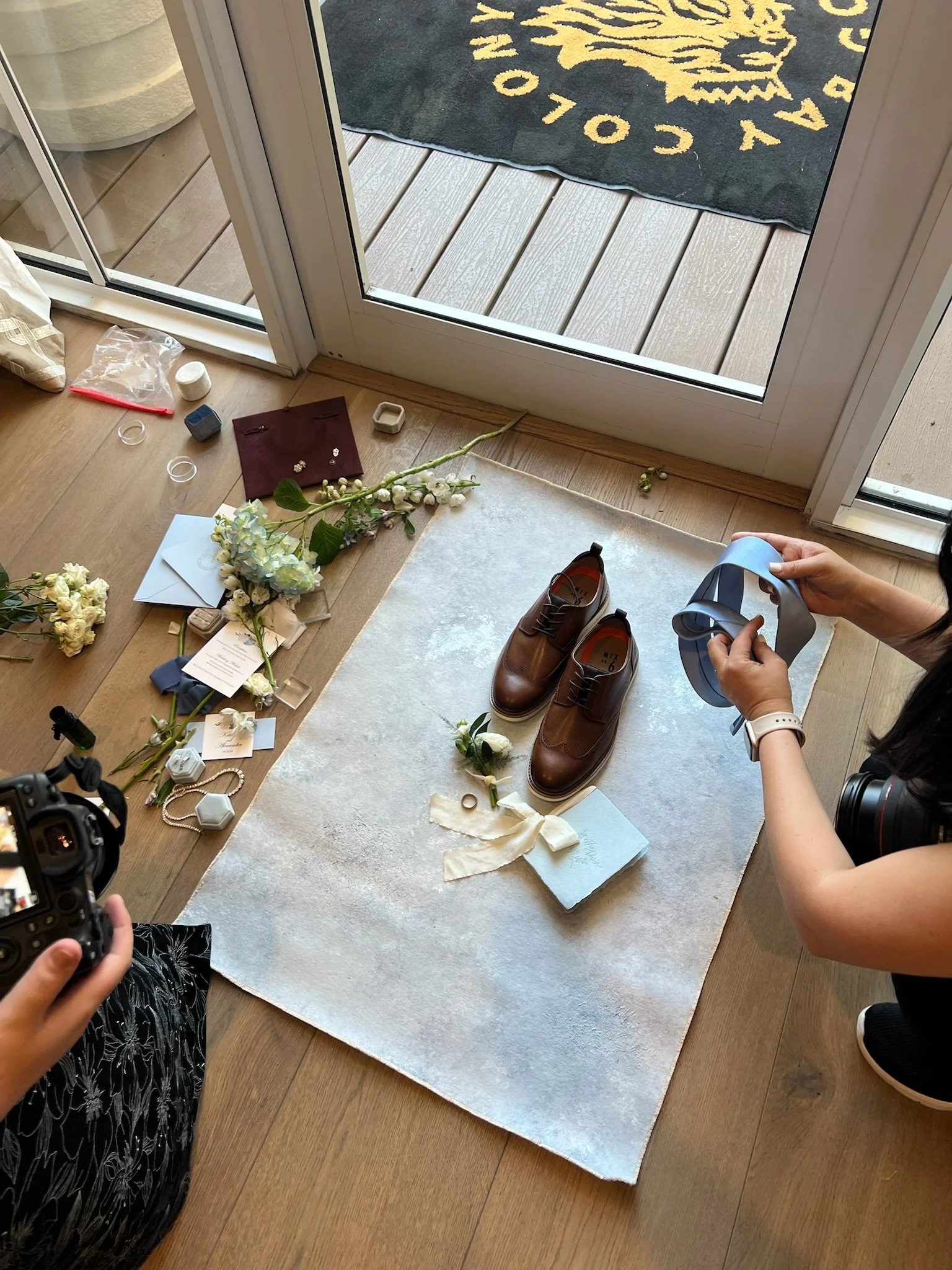View of a door with a welcome mat outside. Inside, a flat surface with various items including floral arrangements, jewelry, a camera, and miscellaneous objects. A person is holding a blue ribbon.