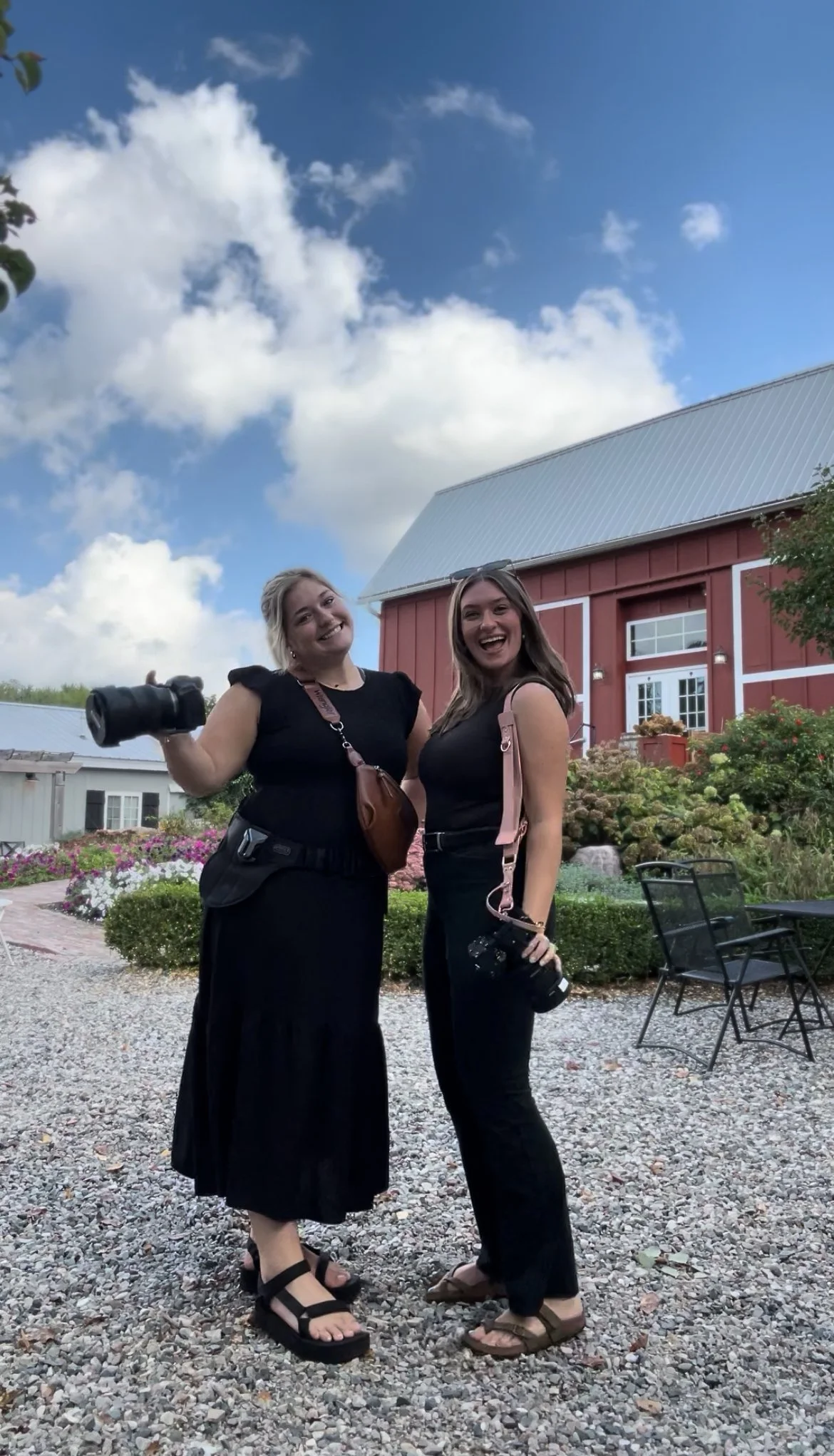 Two women standing outdoors on gravel ground, holding cameras, smiling, with a red barn, flowers, and trees in the background under a partly cloudy blue sky.