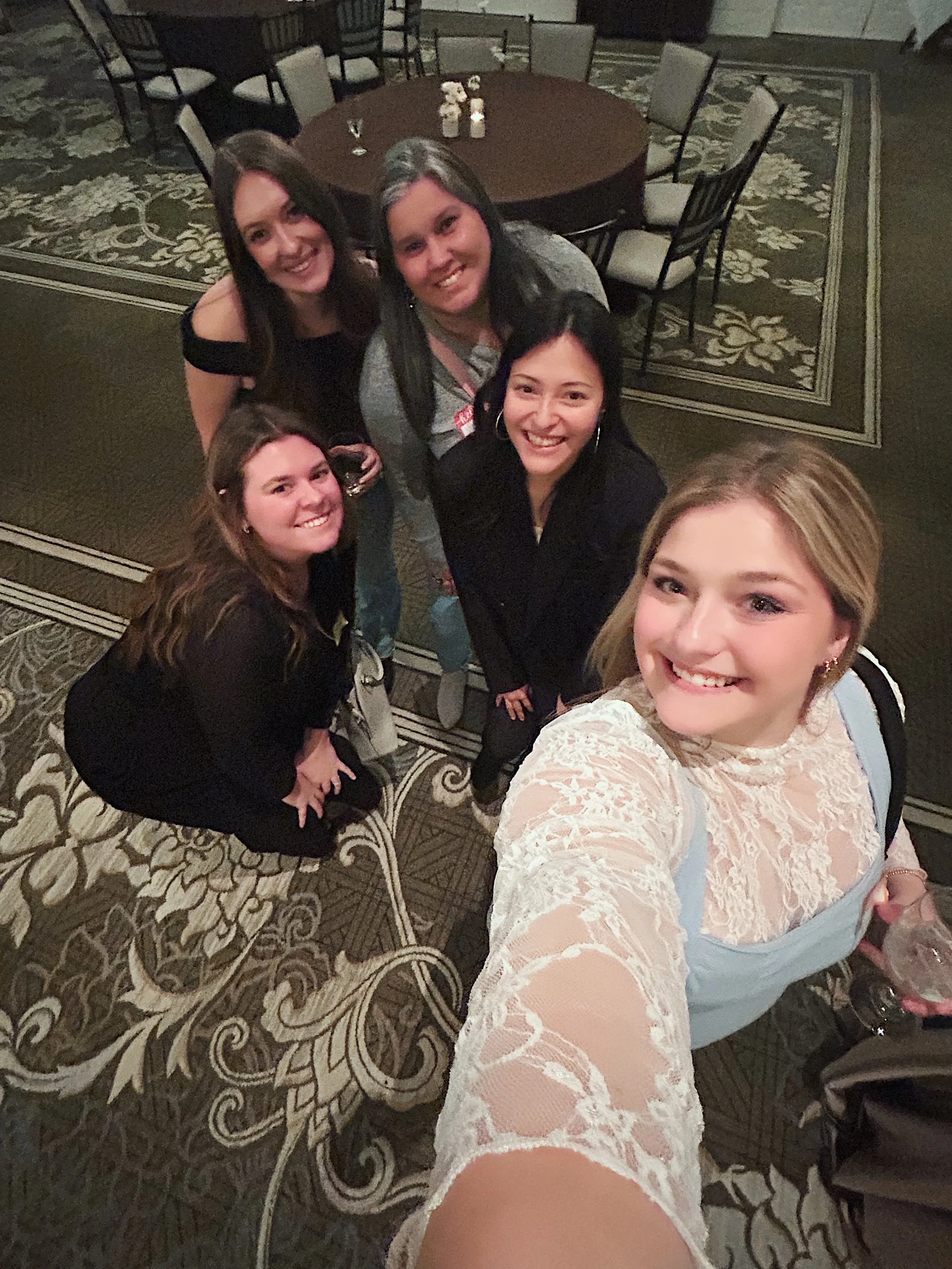 A group of five women taking a selfie at a social gathering or party. They are standing on a decorated carpet near a round table with chairs.