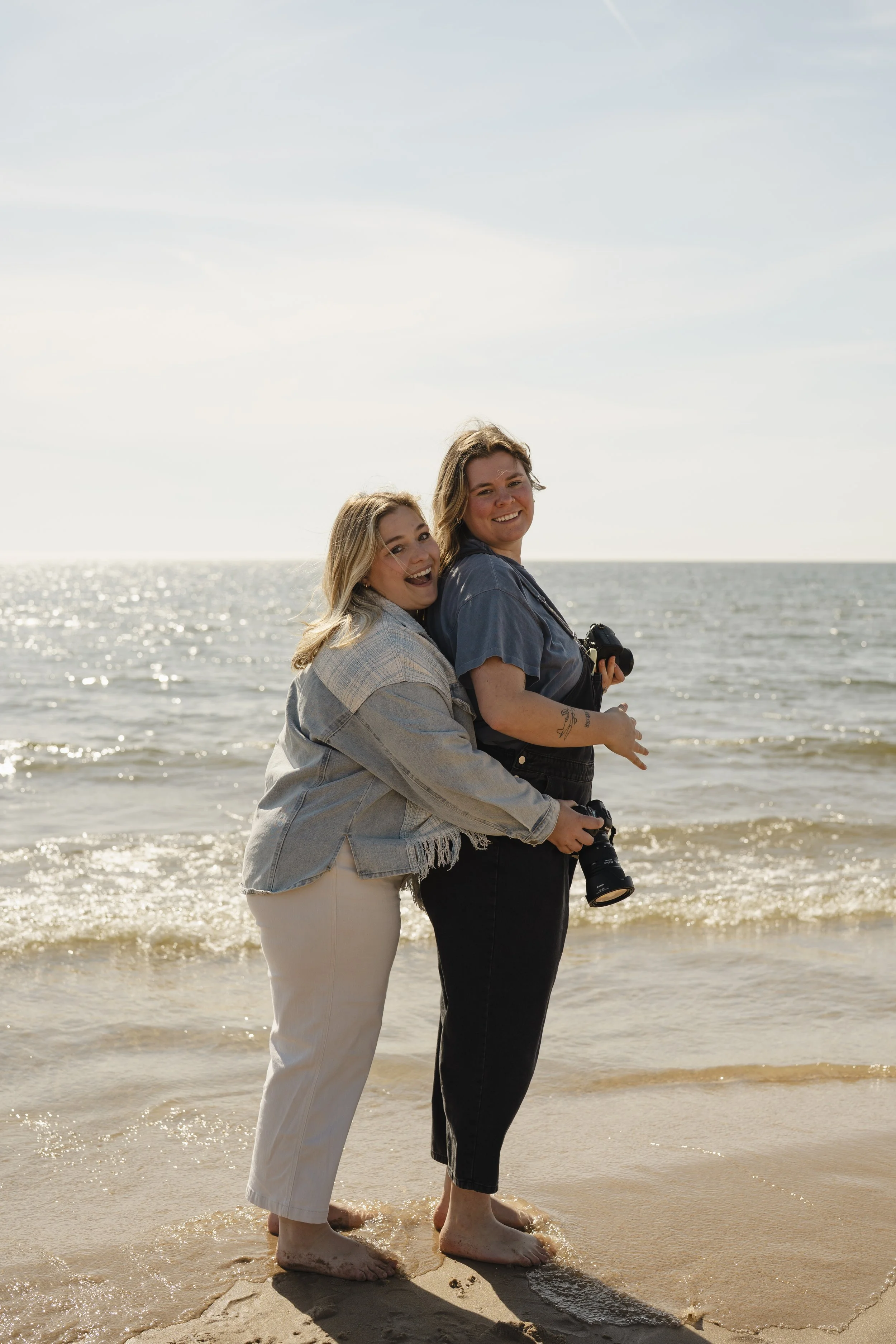 Two women standing on a sandy beach with ocean waves behind them. They are smiling, facing the camera, and leaning against each other. One woman is holding a camera. The sky is partly cloudy.