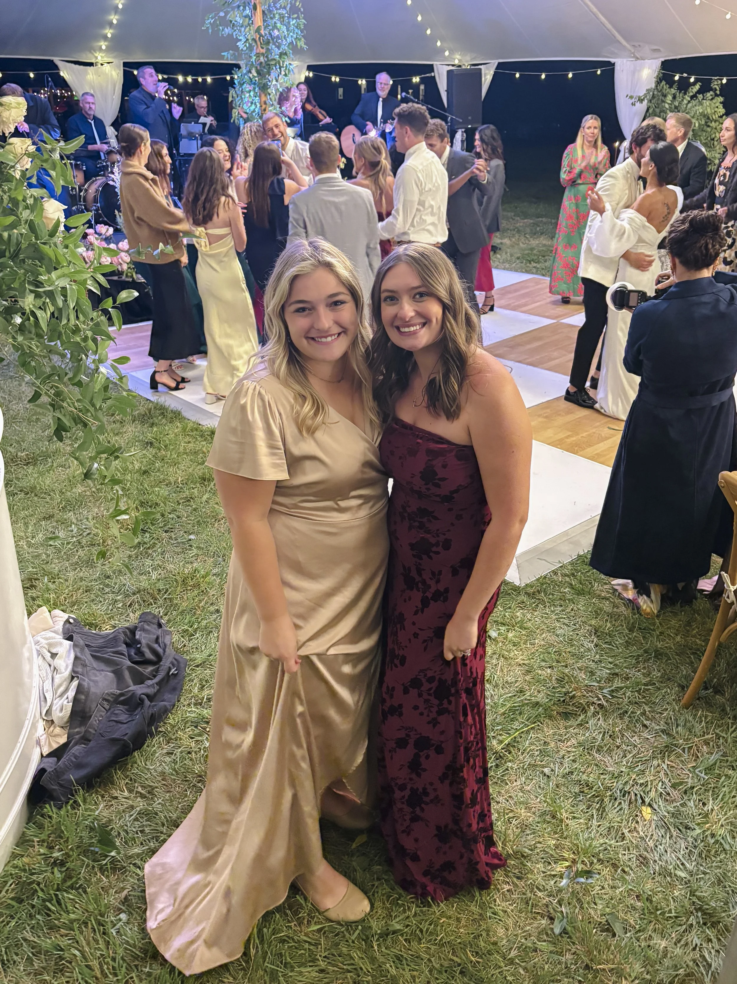 Two smiling women in elegant dresses standing close together at a nighttime celebration under a tent, with people dancing and a live band playing in the background.