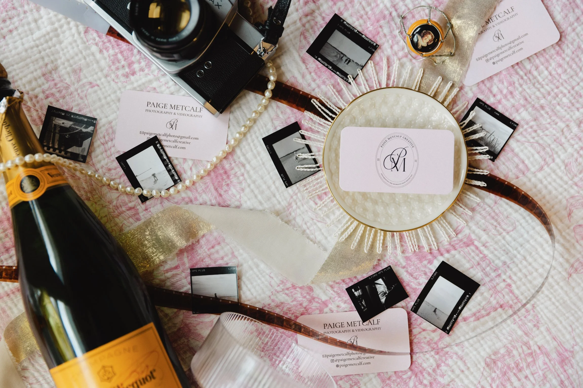 Flat lay of photography and videography items including a camera, business cards, film photographs, a pearl necklace, a bowl with a logo, a bottle of champagne, and gold decorative accents on a pink textured cloth.