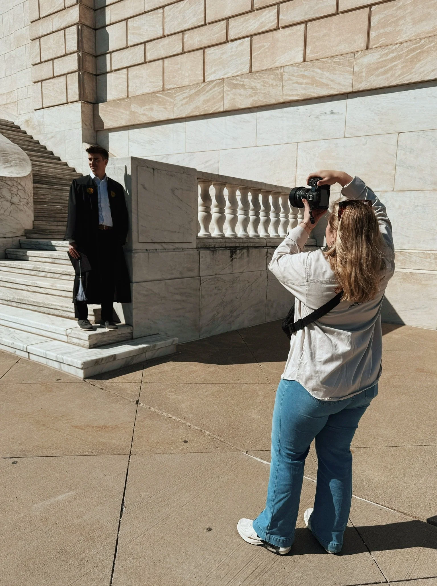 A woman is taking a portrait of a young man in a graduation cap and gown outside a large stone building, with stairs and a railing behind him.