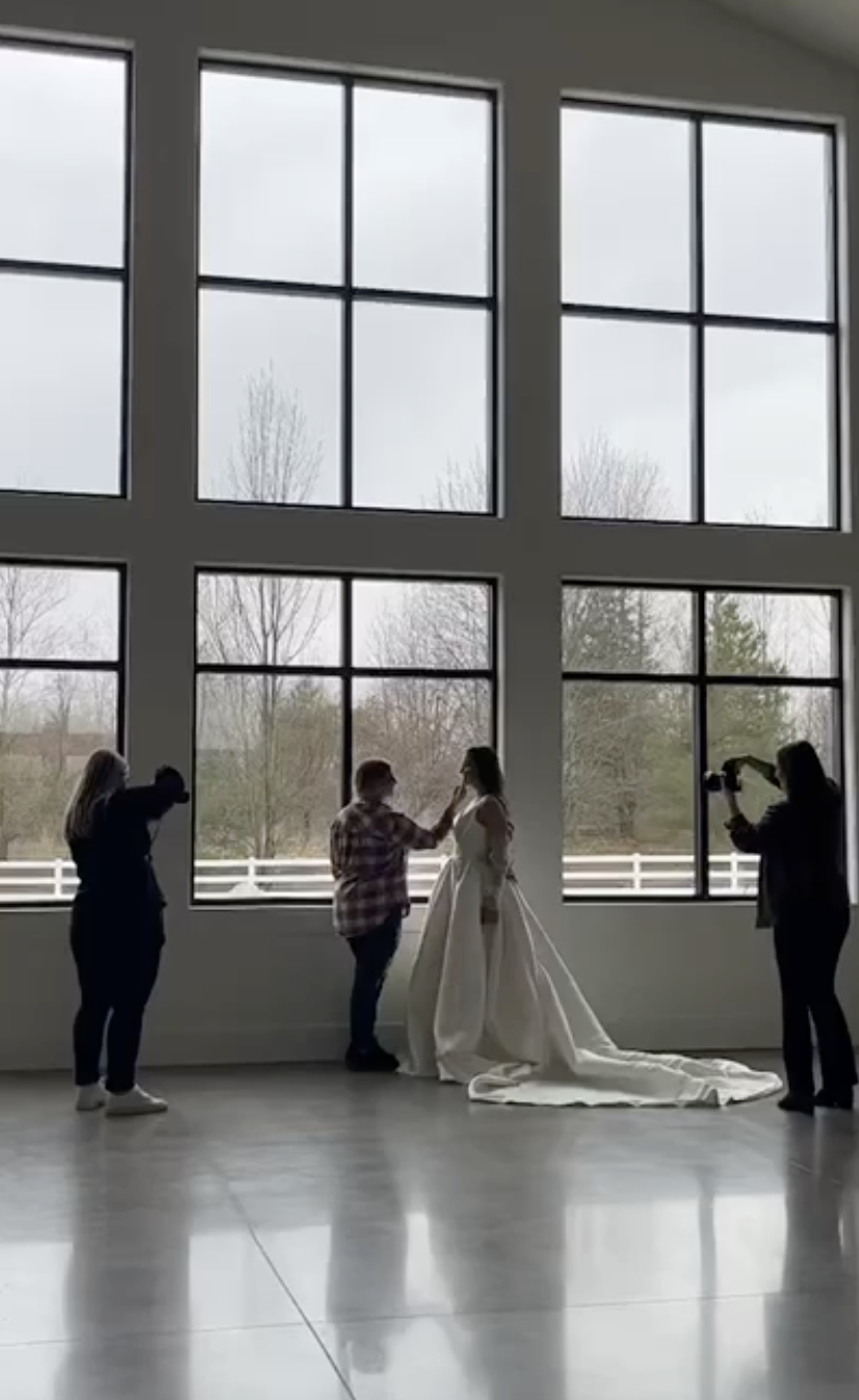 People taking photos of a bride in a white wedding dress standing in front of large window panes.