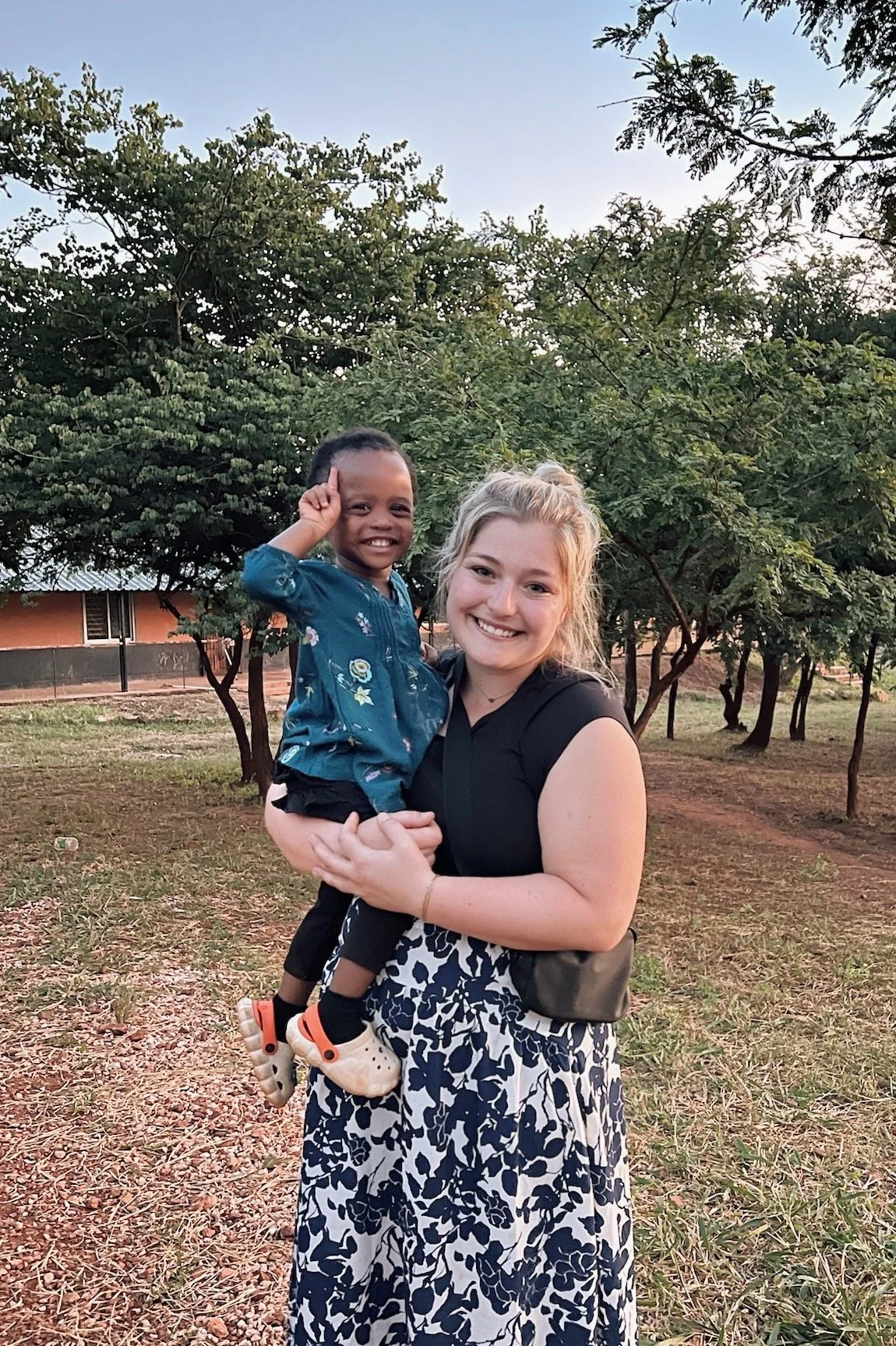 A woman holding a young girl outdoors in a park with trees and a building in the background, both smiling at the camera.
