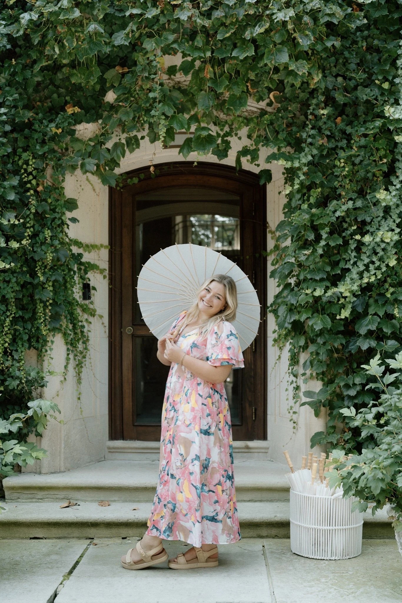 A woman stands on a porch in front of a wooden door surround by green ivy, holding a white parasol, wearing a long floral dress, beige sandals, and smiling.