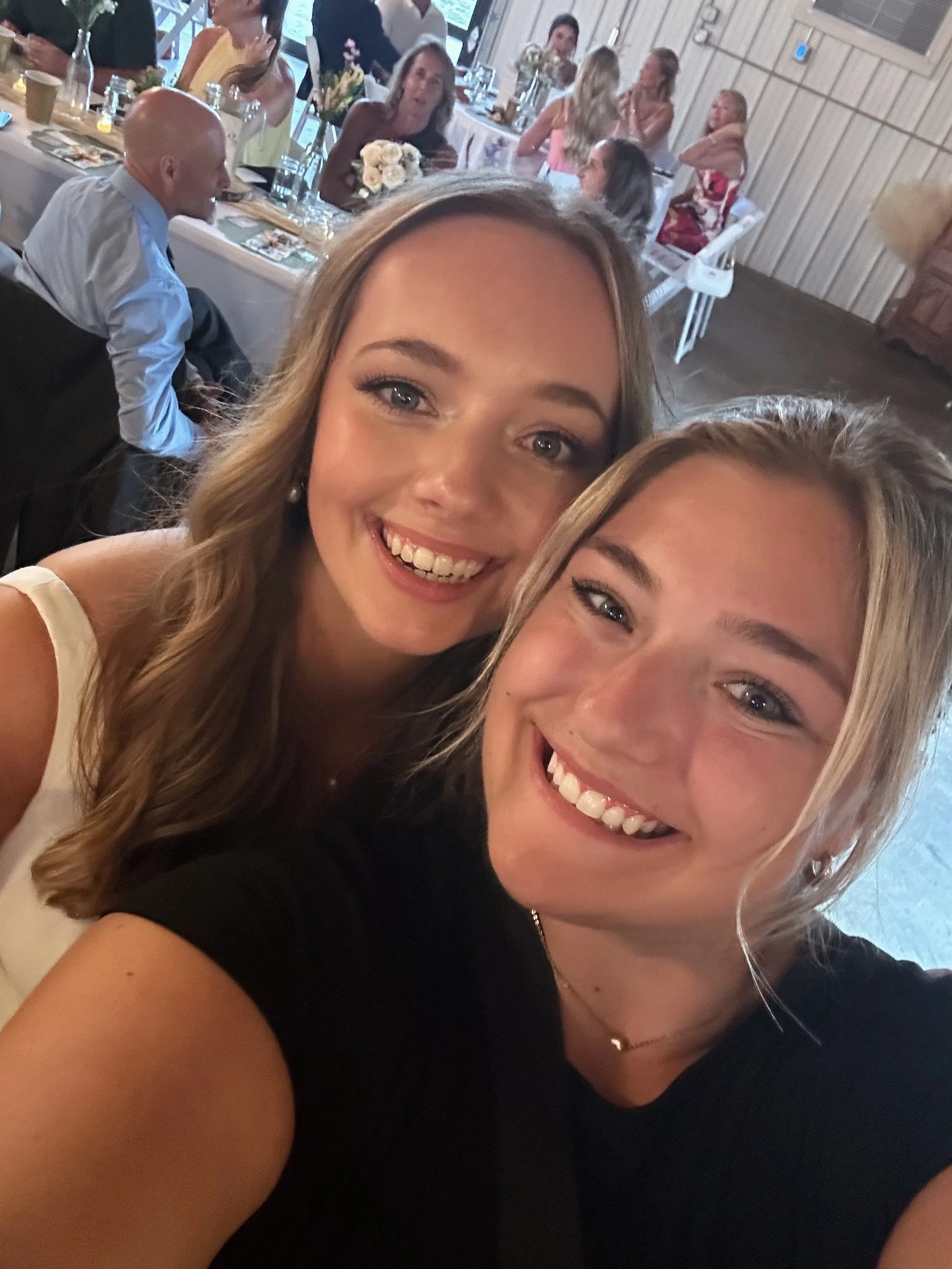 Two women smiling and taking a selfie at a social gathering, with a dinner setup and other guests in the background.