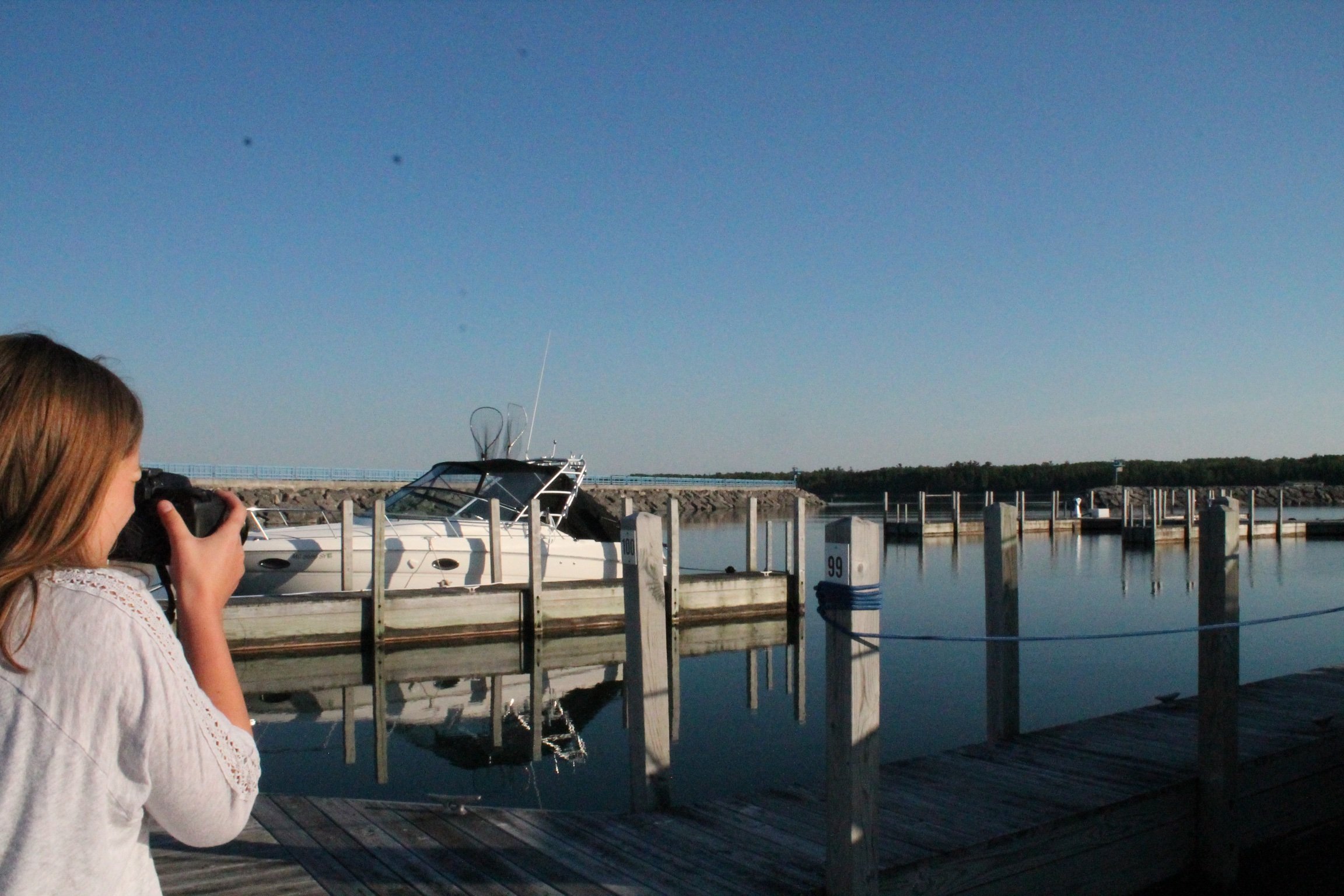 A woman with long brown hair wearing a white blouse is taking a photograph of a boat docked at a marina, with the water, dock, and clear blue sky visible in the background.