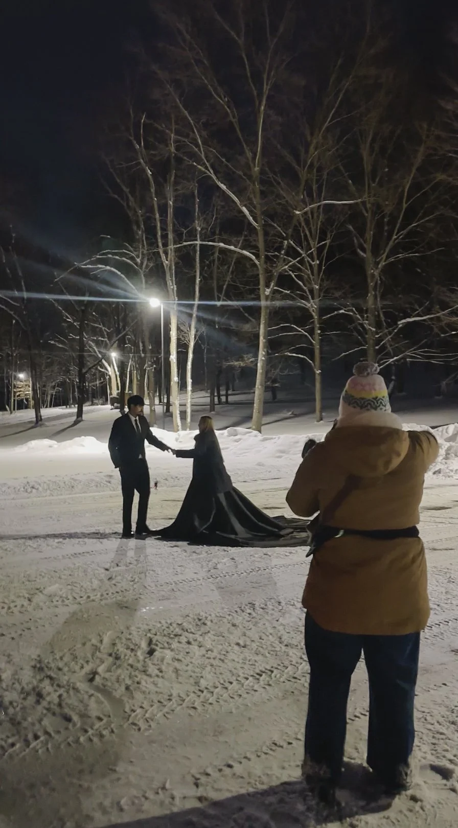 A couple dressed in formal attire, holding hands and standing in the snow at night in a park, with a woman taking photos of them, in a winter setting with bare trees and snow-covered ground.