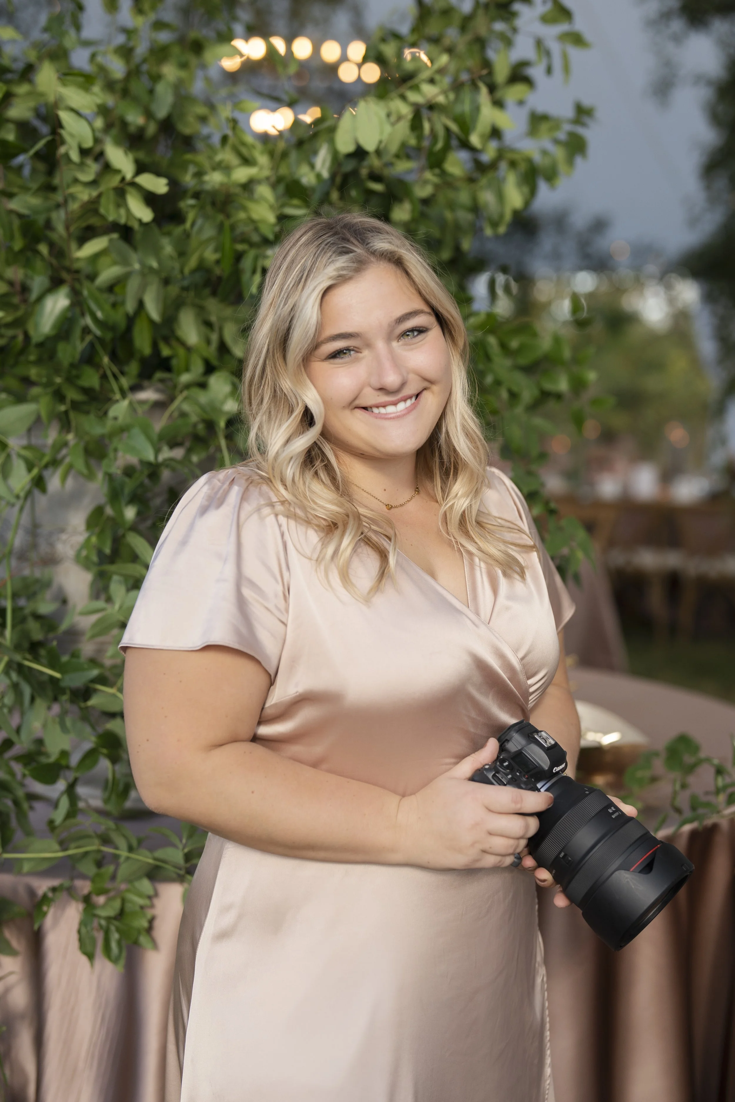 A young woman with blonde, wavy hair, smiling, holding a professional camera outdoors during the evening. She is wearing a beige satin dress and a delicate gold necklace, with green foliage and blurred lights in the background.