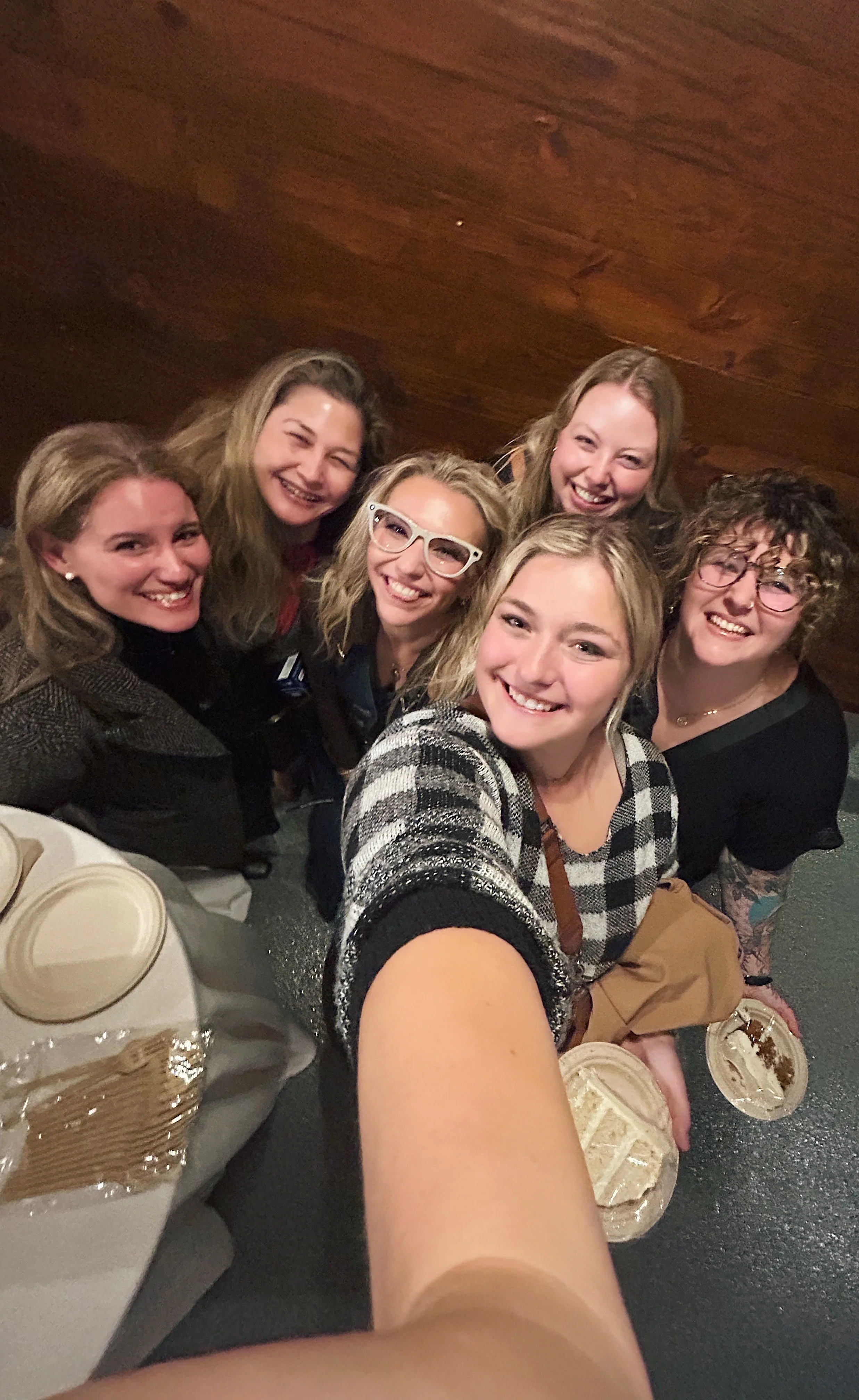 Group of six women smiling and taking a selfie at a social gathering, with dining tables and desserts in the foreground.