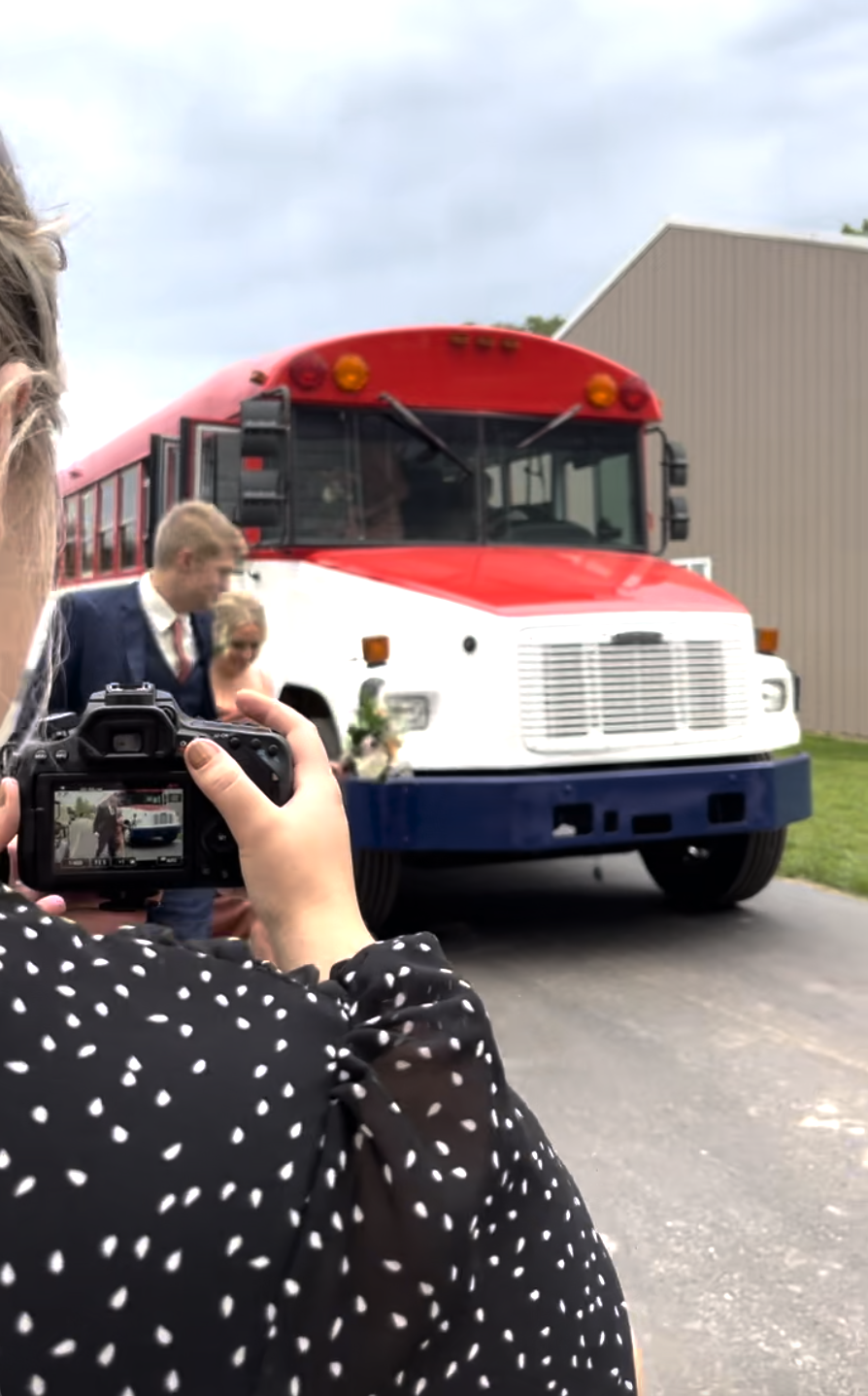 People at a wedding or event taking pictures of a red, white, and blue bus with a person holding a camera in the foreground