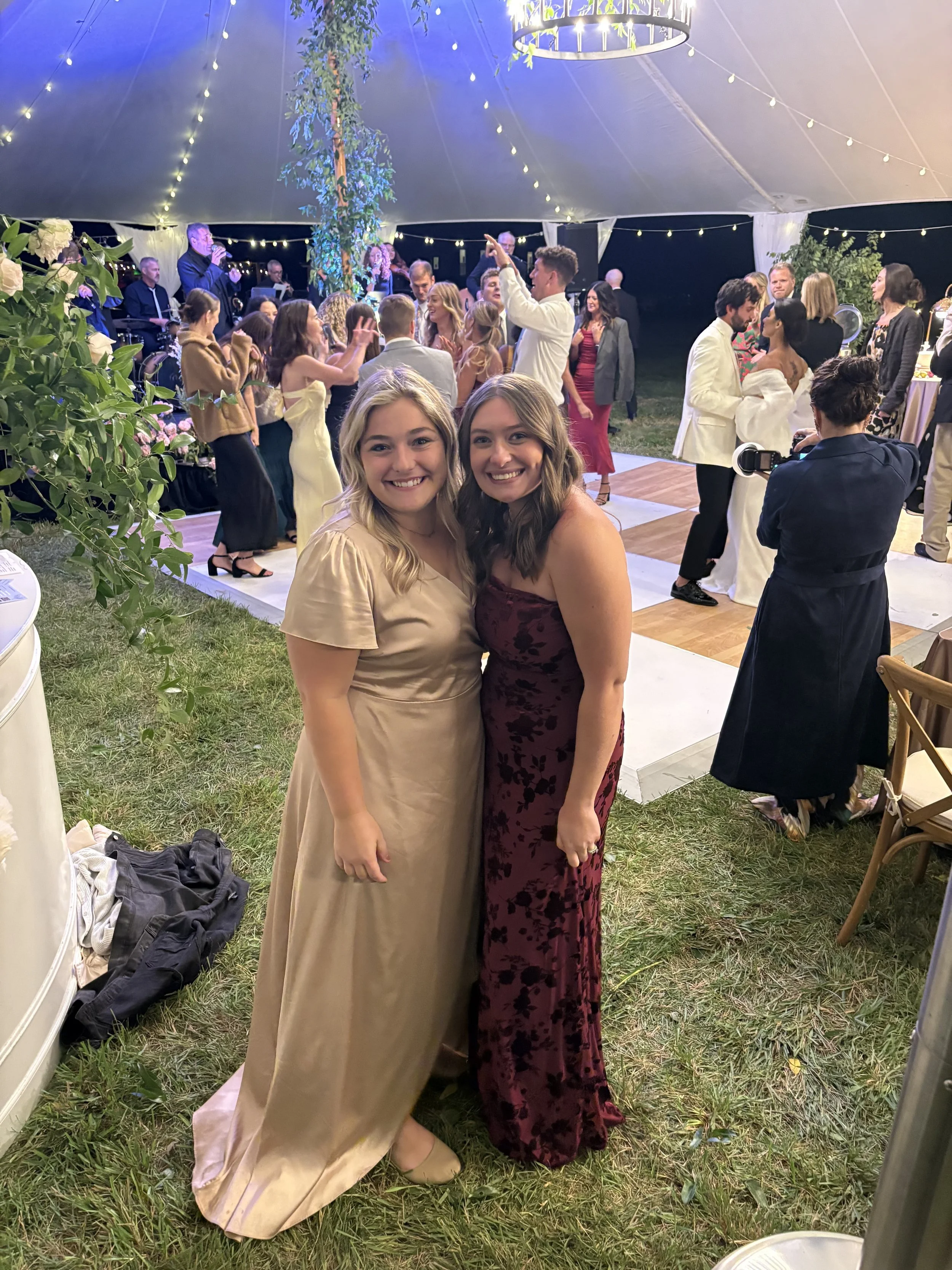 Two women standing together at a nighttime outdoor celebration under a tent, smiling at the camera. They are dressed elegantly, with one in a cream-colored gown and the other in a dark red floral dress. The background shows dancing guests, musical ba