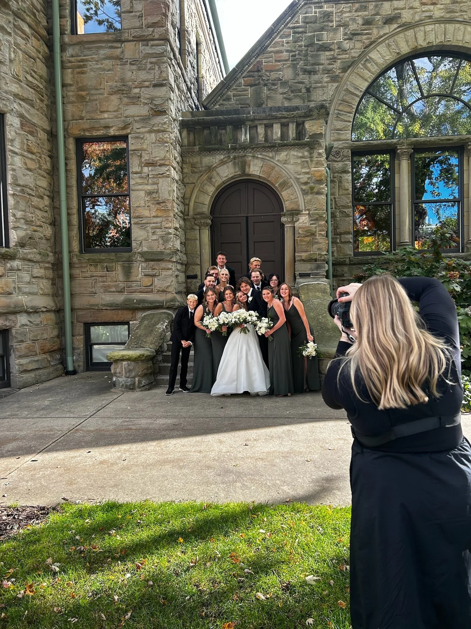 A group of people in formal attire, some holding flowers, are gathered outside in front of a stone building with large windows, posing for a photograph taken by another person.