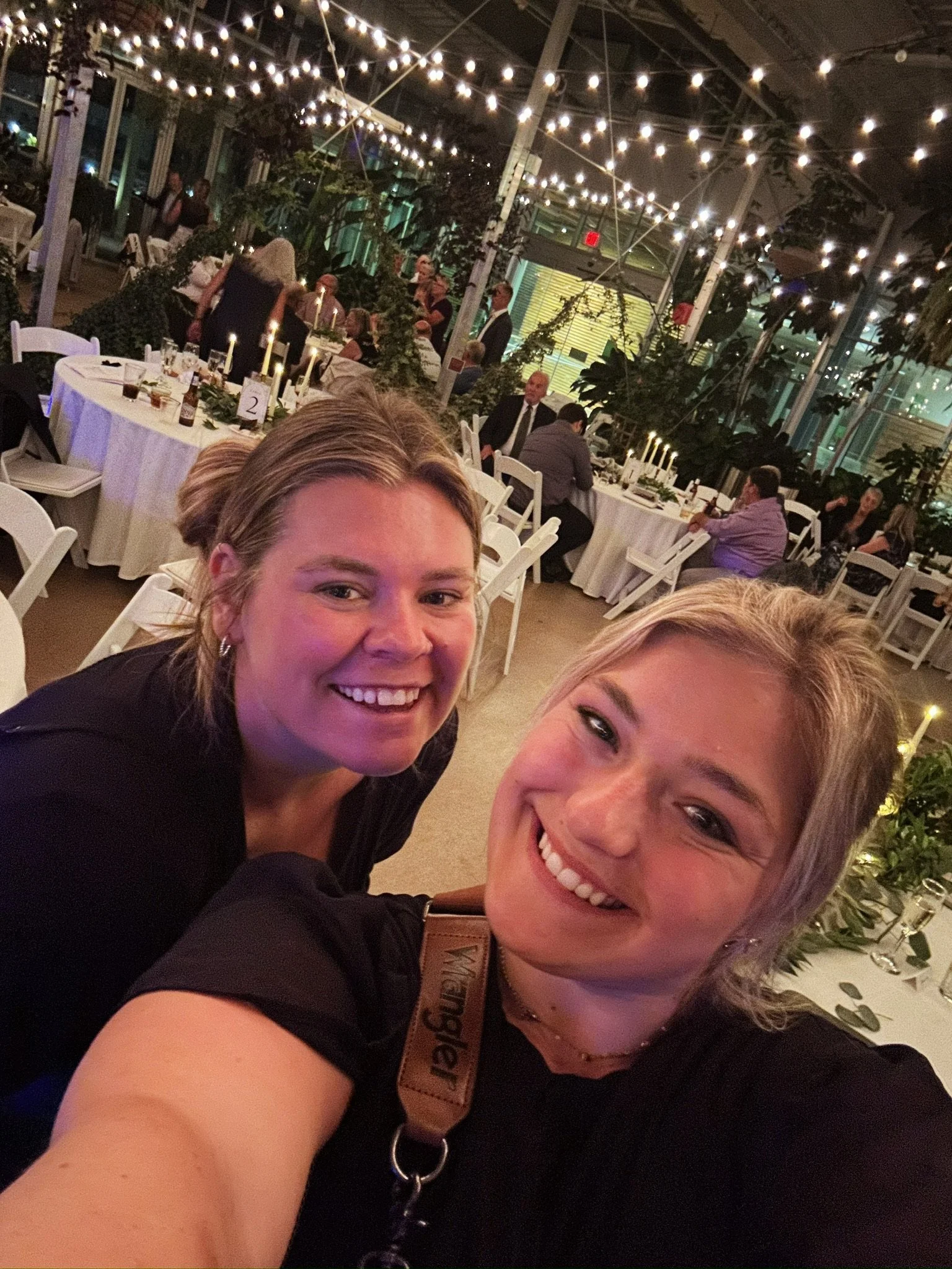 Two smiling women taking a selfie at a festive indoor event with string lights, tables, and people in the background.