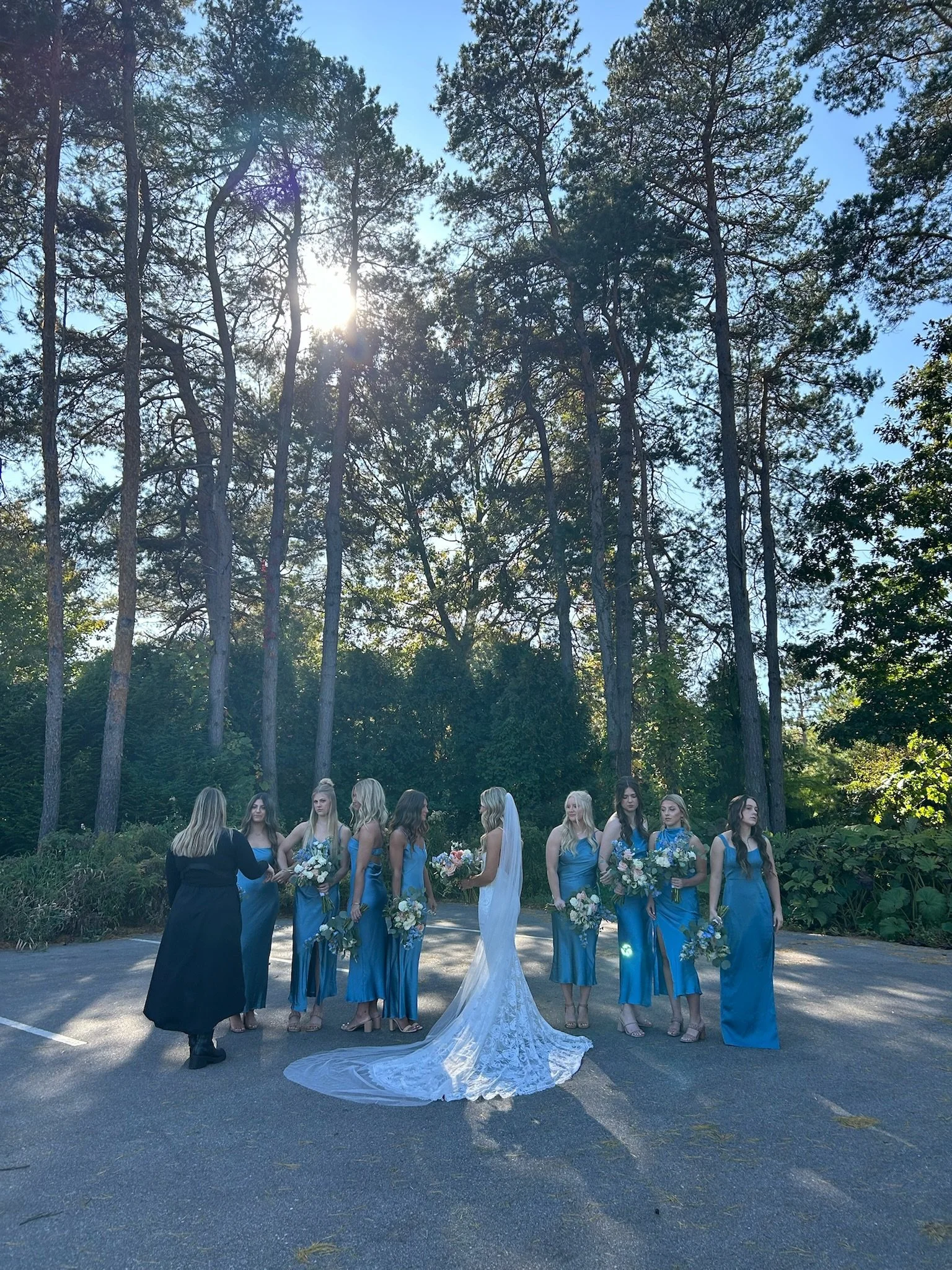 Bridal party of nine women in blue dresses standing outdoors, with a bride in a white gown and veil, holding bouquets of flowers, in front of tall trees and sunlight.
