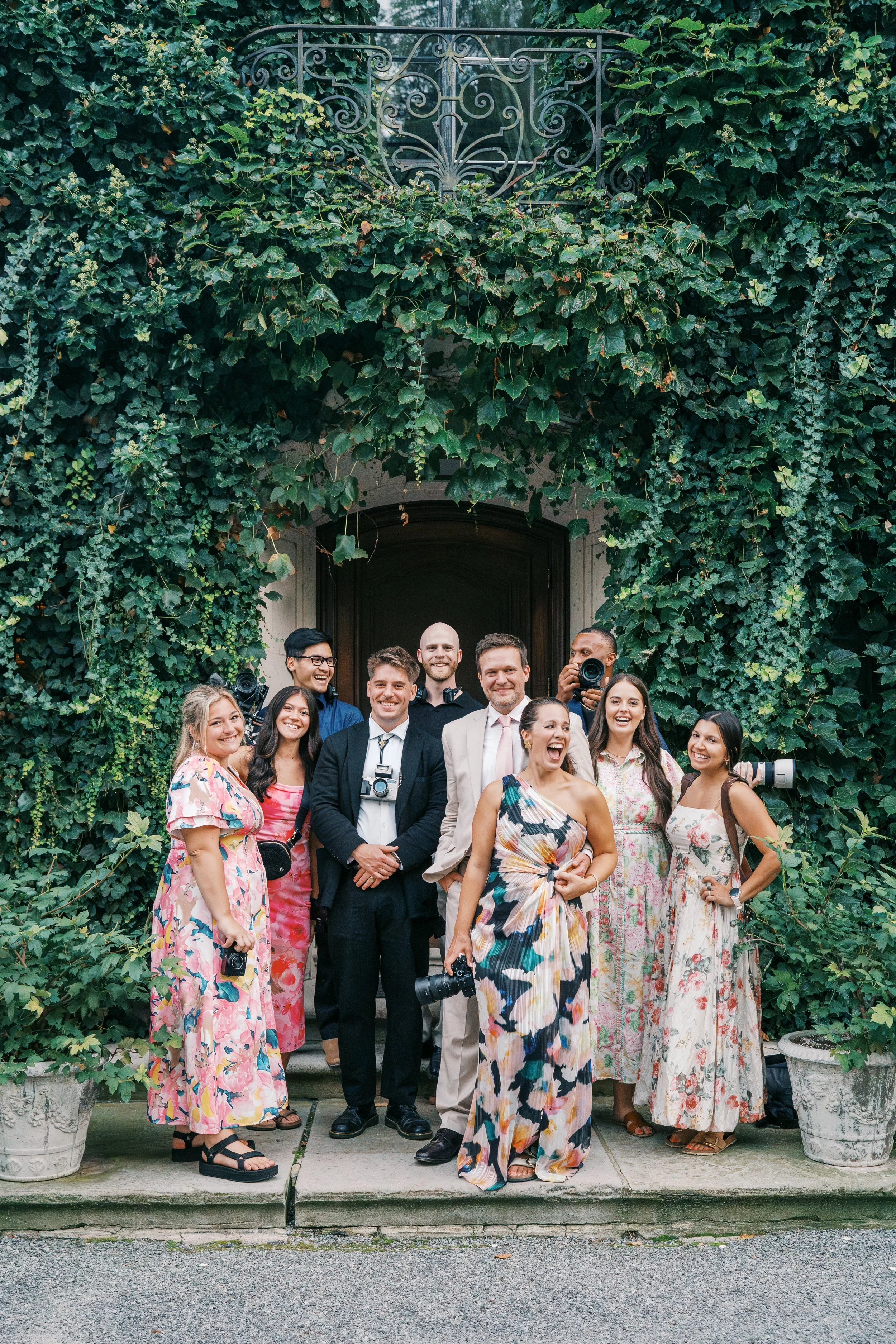 Group of diverse people smiling and standing together outside in front of a building with a large wooden door and green ivy-covered walls. The group includes men and women, some holding cameras.