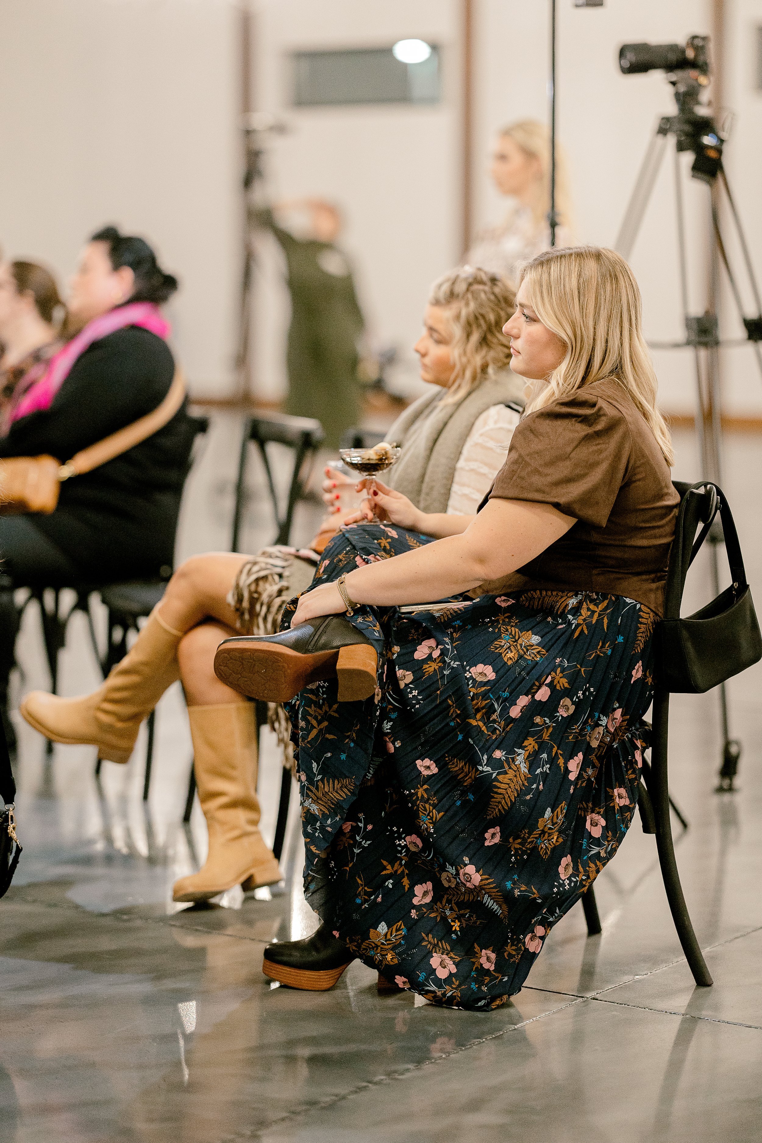 A group of women sitting on chairs at an event, with some holding drinks, in an indoor setting with a camera on a tripod in the background.