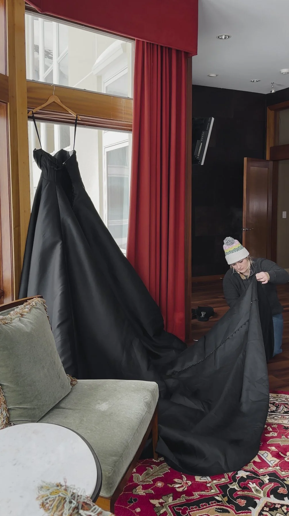 A woman wearing a wool hat and dark jacket is folding a black fabric, likely a gown, in a room with wooden furniture and red curtains. A dress or gown is hanging on a wooden hanger near a window.