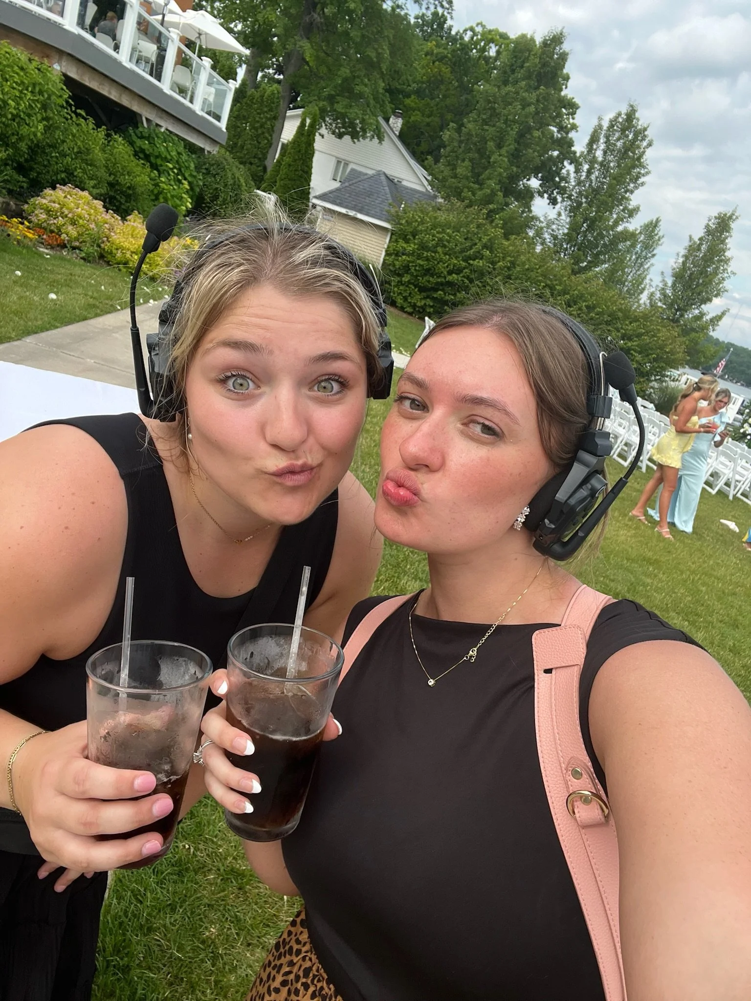 Two women taking a selfie at an outdoor event, each holding a glass of dark soda with ice. They are wearing headsets, and there are people and white chairs in the background.