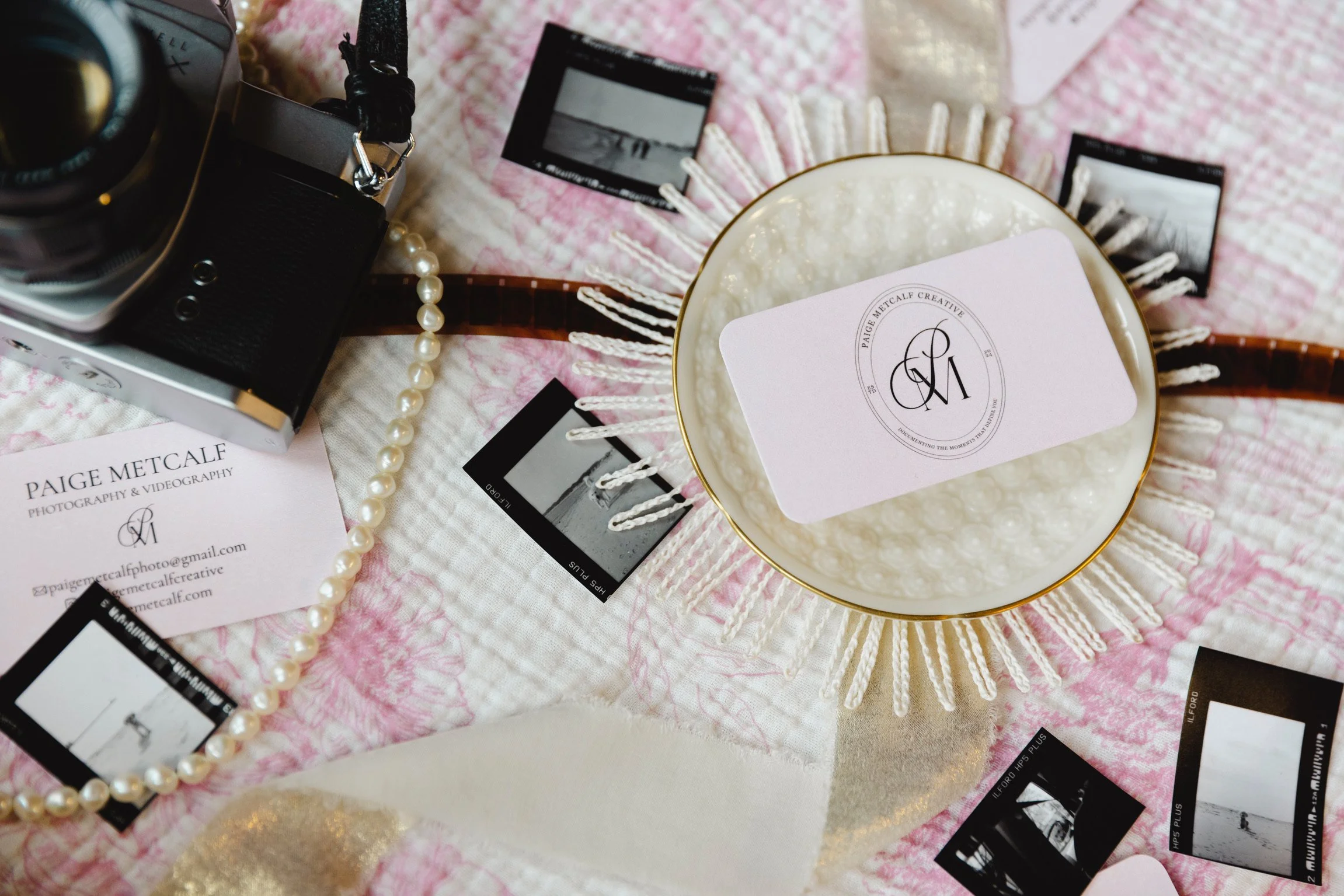 A flat lay of photography and videography equipment on a pink and white textured cloth, including a camera, film slides, pearl necklace, a decorative bowl with a business card, and a fringed hanging decoration.