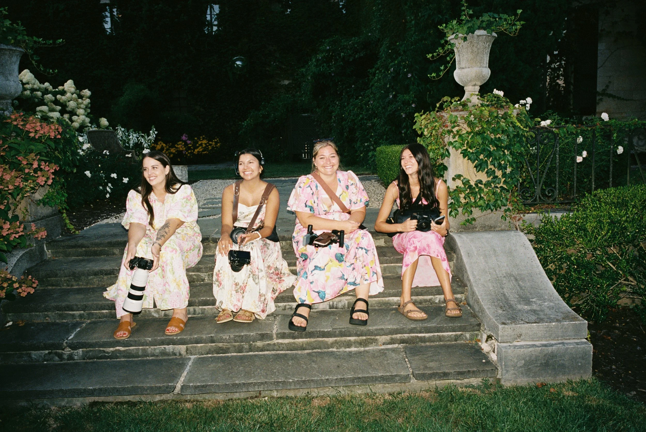 Four women sitting on stone steps in a garden with lush greenery and flower pots, smiling and holding cameras, dressed in floral summer dresses.
