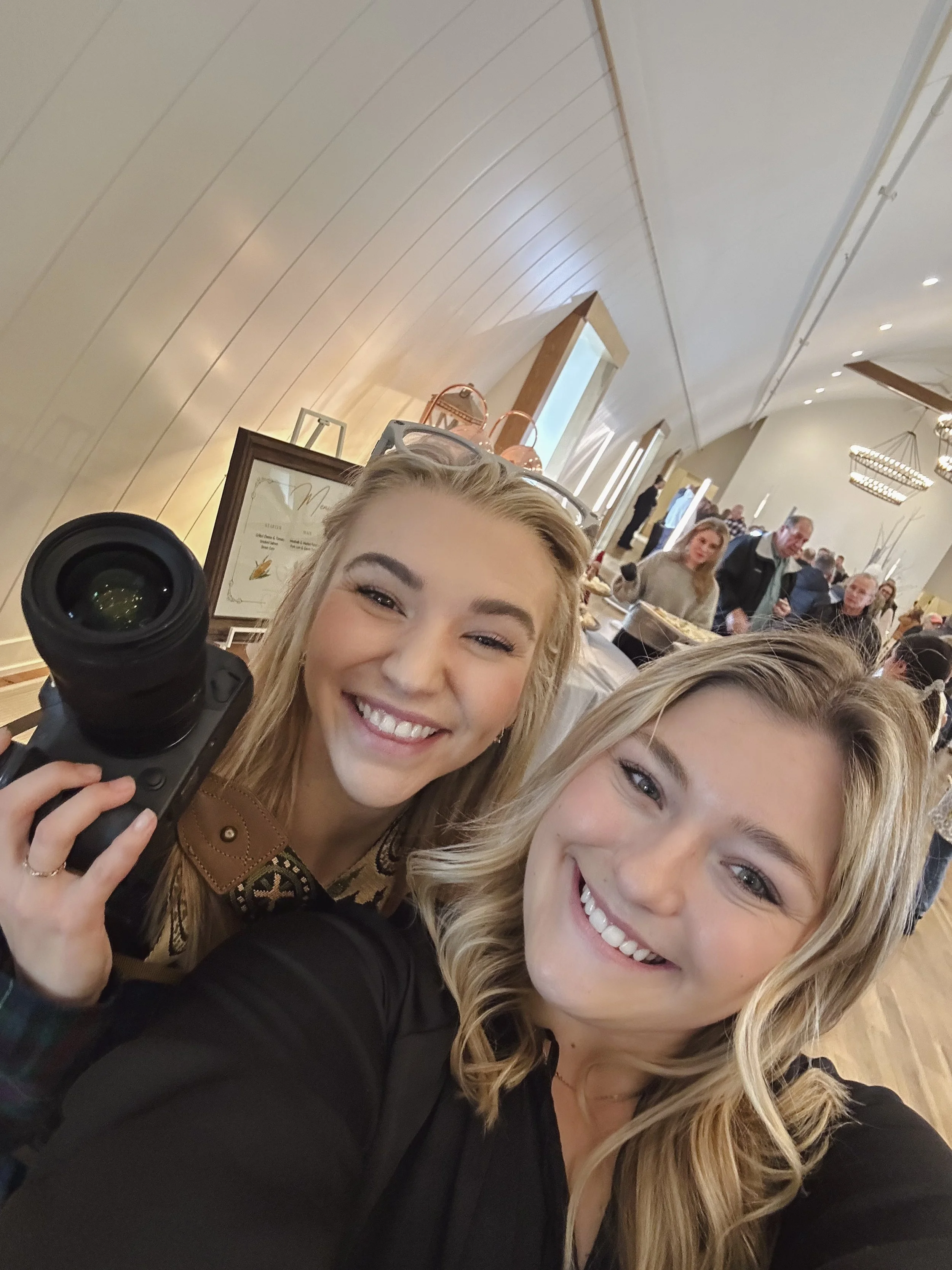 Two smiling blonde women taking a selfie in a crowded indoor setting, with multiple people in the background, some at a counter, in a warmly lit room with chandelier lighting and window details.