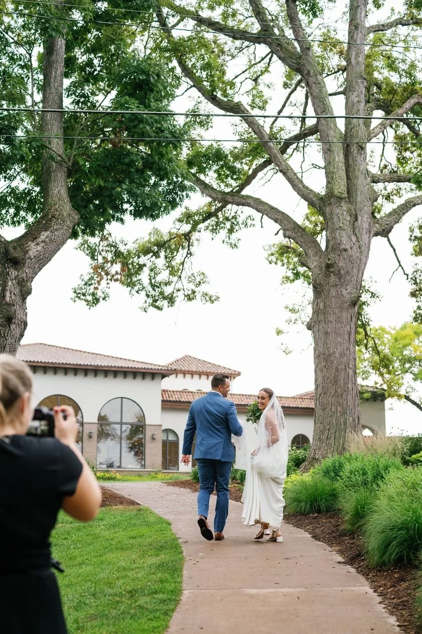 A bride and groom walking on a sidewalk outside a building, smiling at each other, with a photographer capturing the moment.