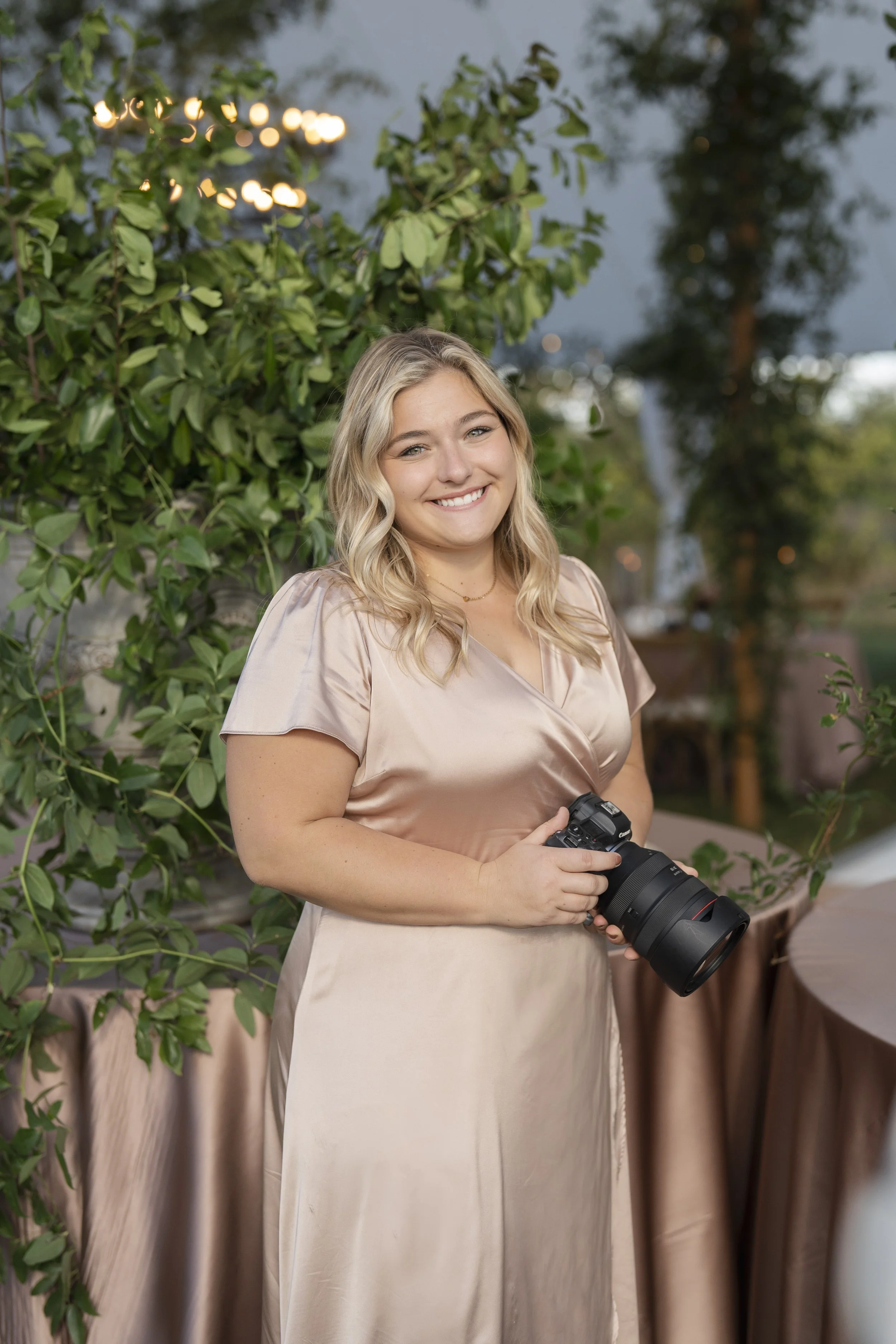 A young woman with blonde curly hair smiling while holding a professional camera with a large lens. She is wearing a shiny beige dress and standing outdoors in front of lush green plants and trees during the evening.