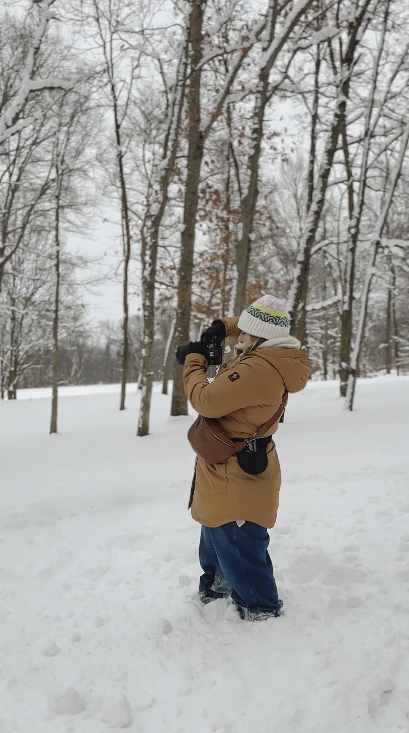 A person dressed warmly in a tan winter coat, blue jeans, and a knit hat with colorful patterns takes a photo with a camera in a snowy, leafless forest.