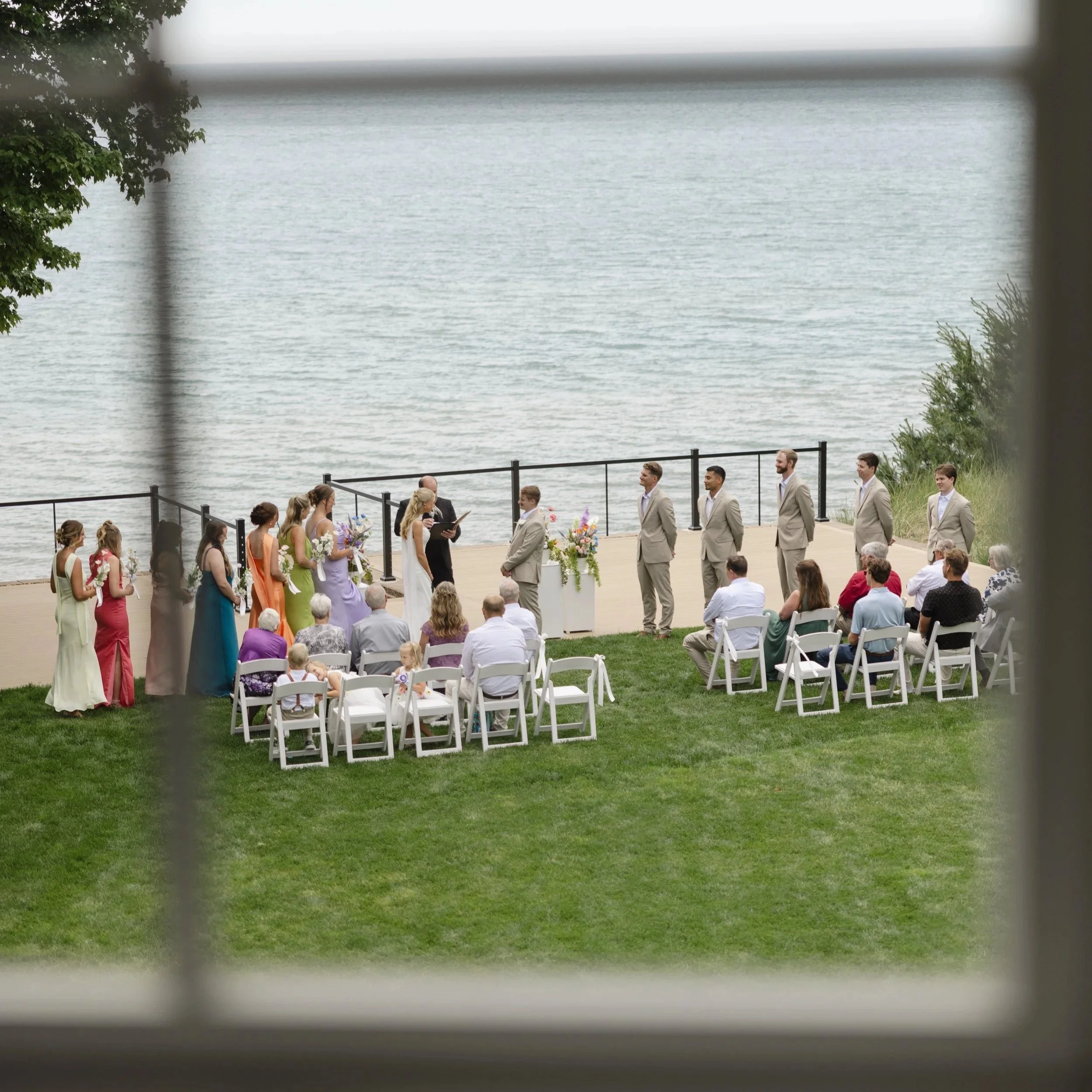 A wedding ceremony taking place outdoors by a body of water, with guests seated, the bride and groom standing, and officiant present, surrounded by greenery.