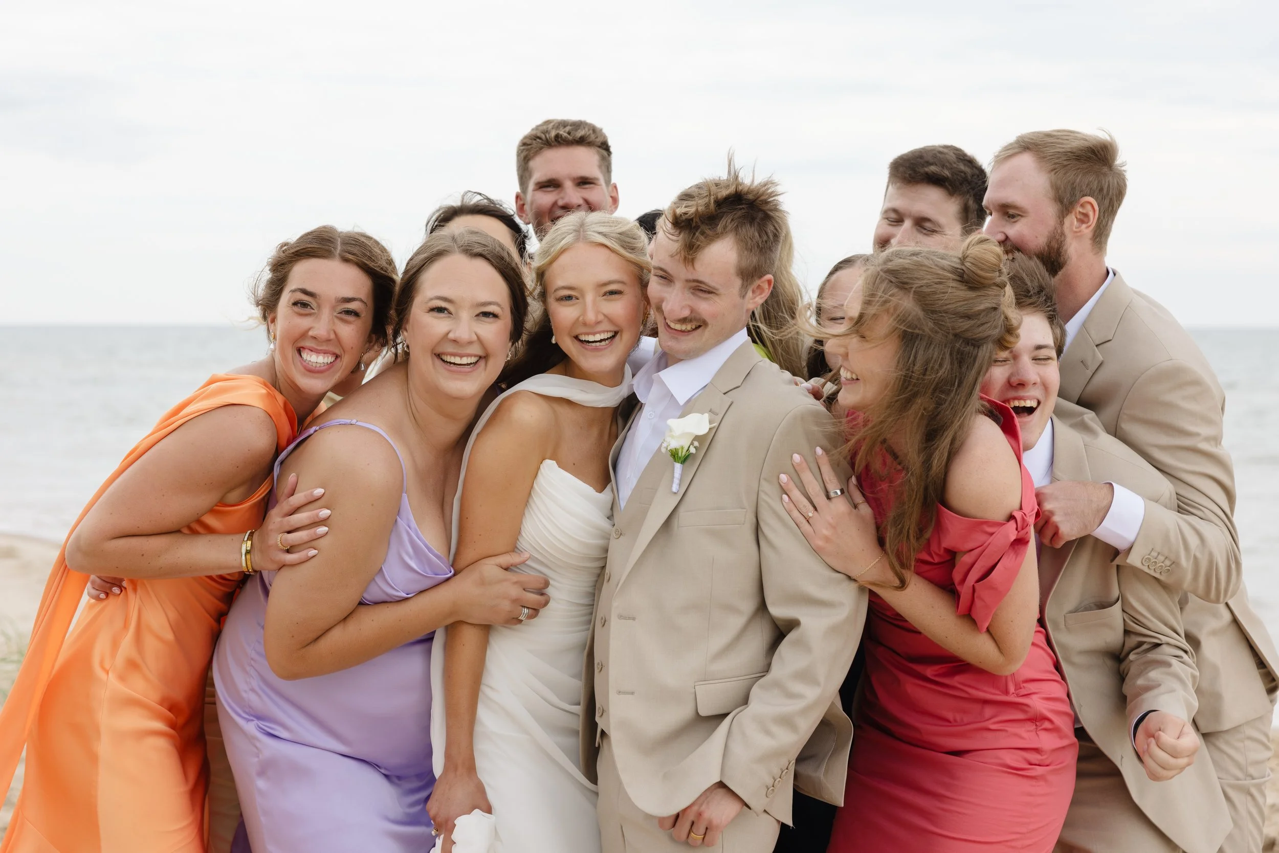 Group of friends at a wedding on the beach, wearing colorful dresses and beige suits, smiling and hugging.