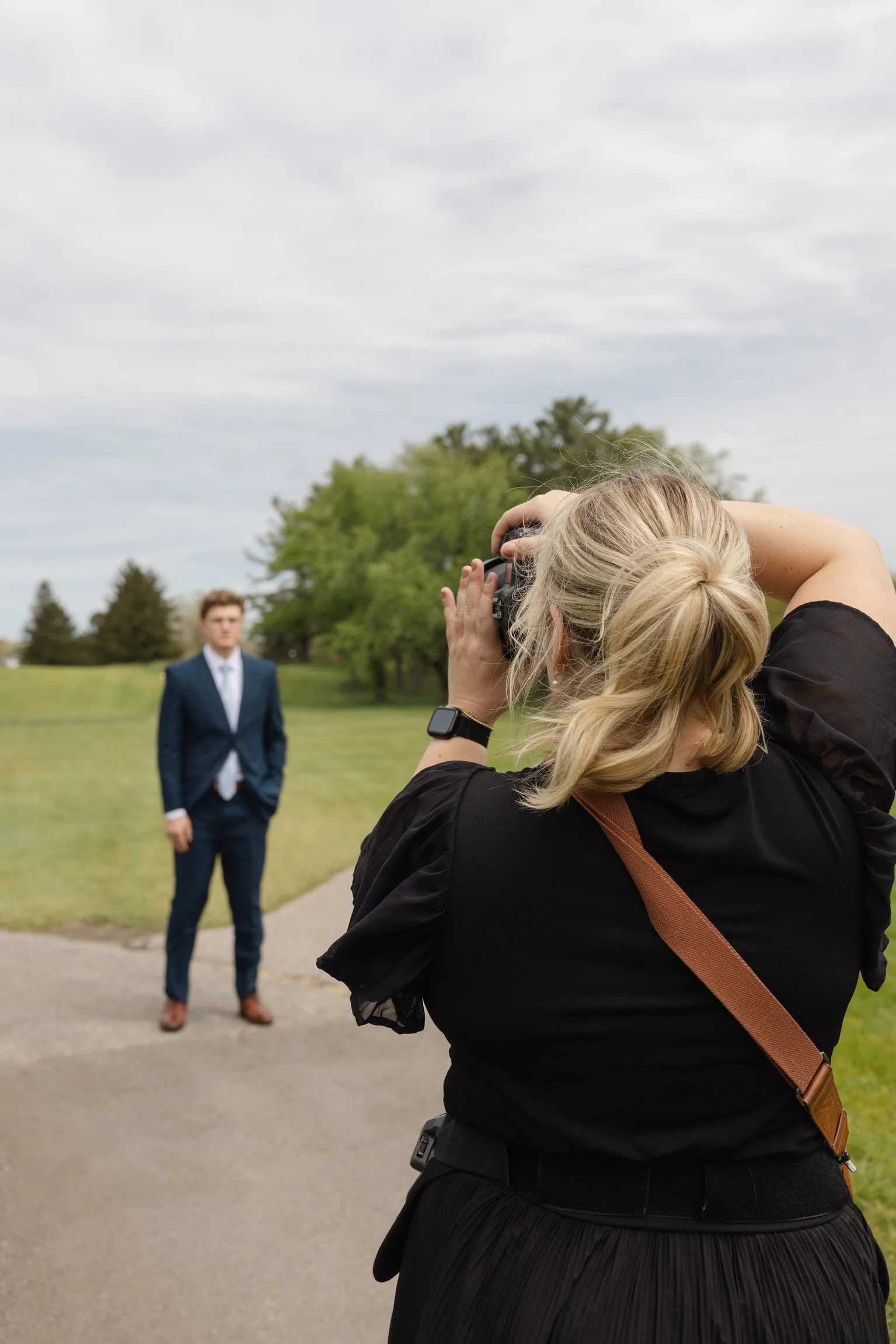 Photographer taking a picture of a man in a navy blue suit with a white shirt and brown shoes in a park with trees and cloudy sky.