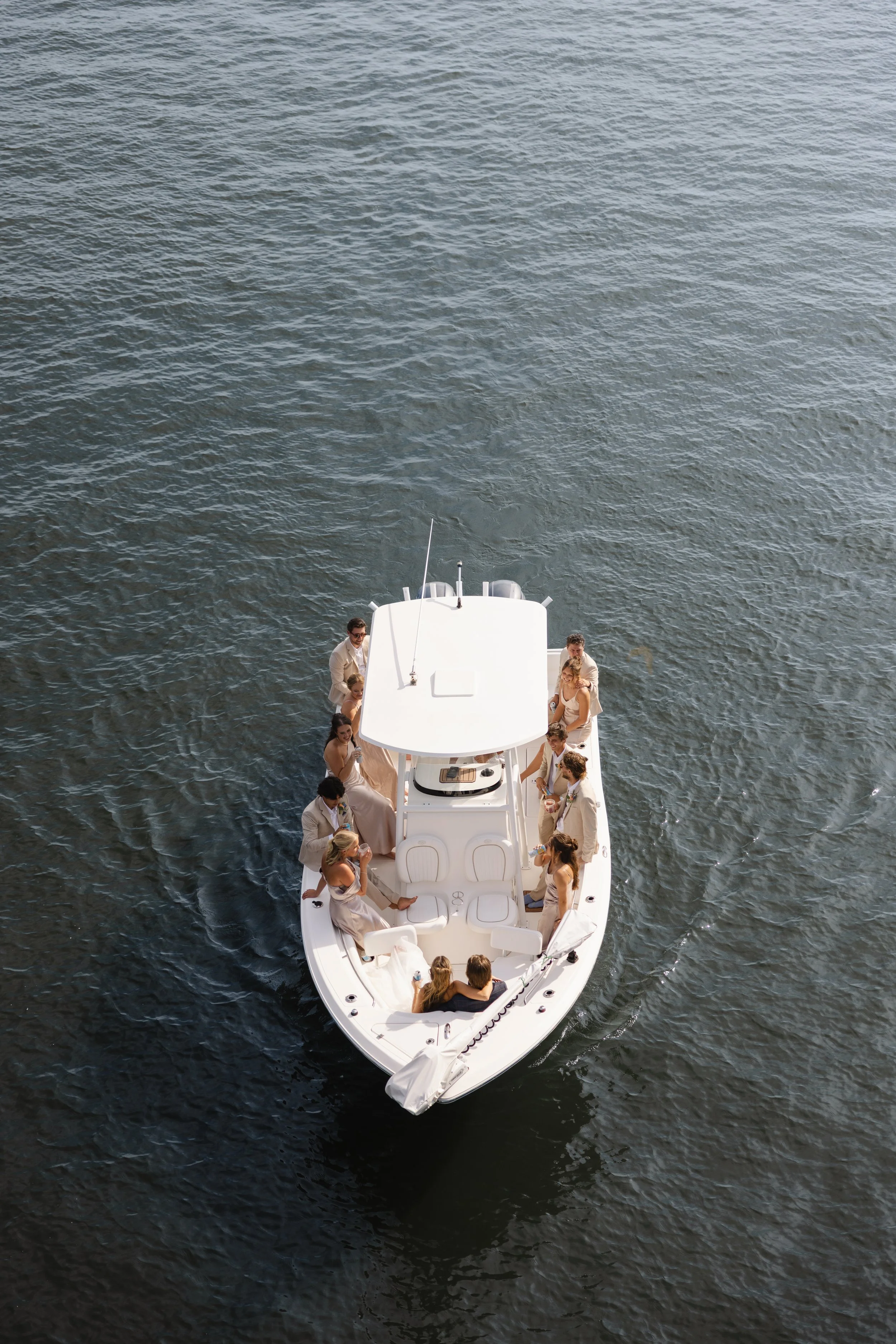 An aerial view of a white yacht with multiple guests on board, mostly women in formal dresses, floating on dark water.