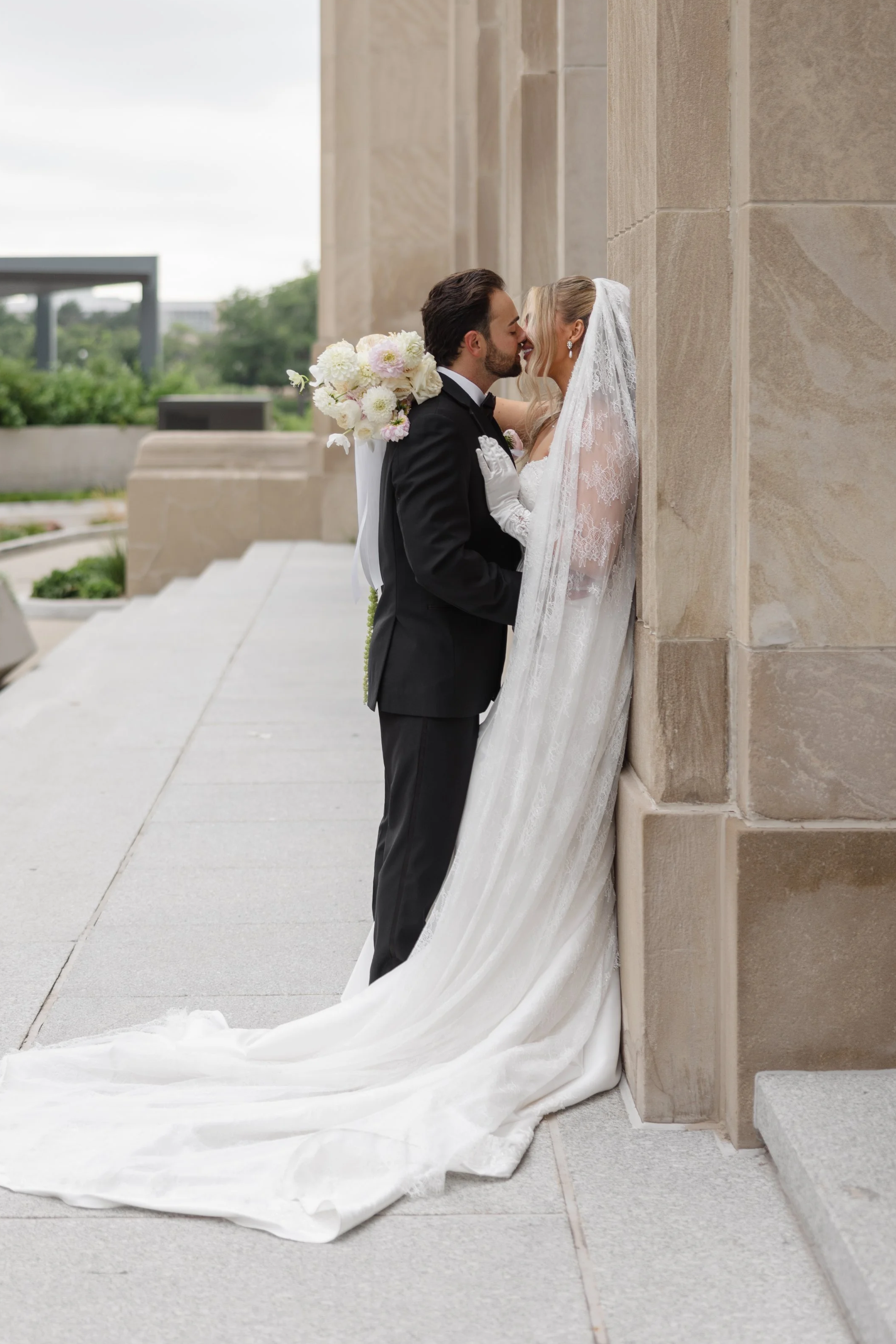 A bride and groom are sharing a kiss outside a stone building. The bride is wearing a lace wedding gown and veil, and the groom is dressed in a black tuxedo. The bride is holding a bouquet of white and light pink flowers.