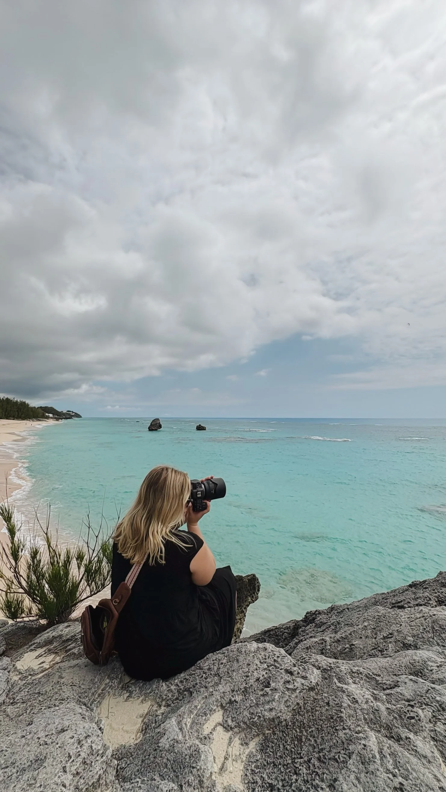 Woman sitting on rocks by the beach taking a photo of the ocean with a camera, cloudy sky overhead