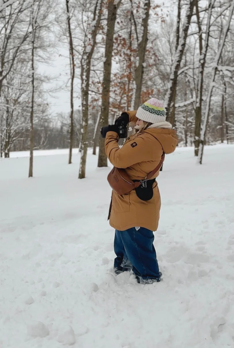 Person in winter clothing taking a photo with a camera in a snowy forest.