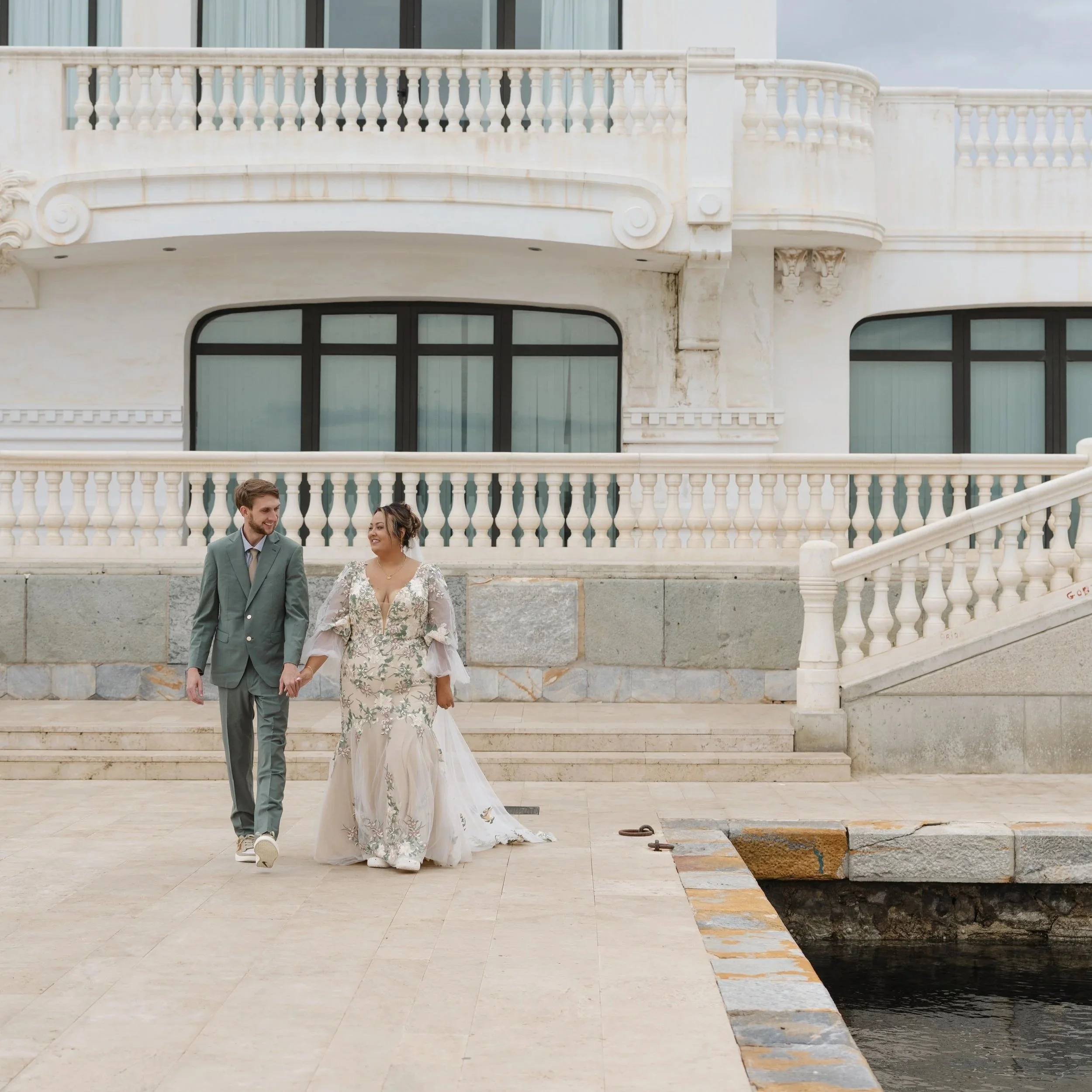 A young couple with light skin and dark hair walking down steps outside a white building with balustrades. The man has a beard and is wearing a gray suit, while the woman is wearing a white floral dress with lace details.