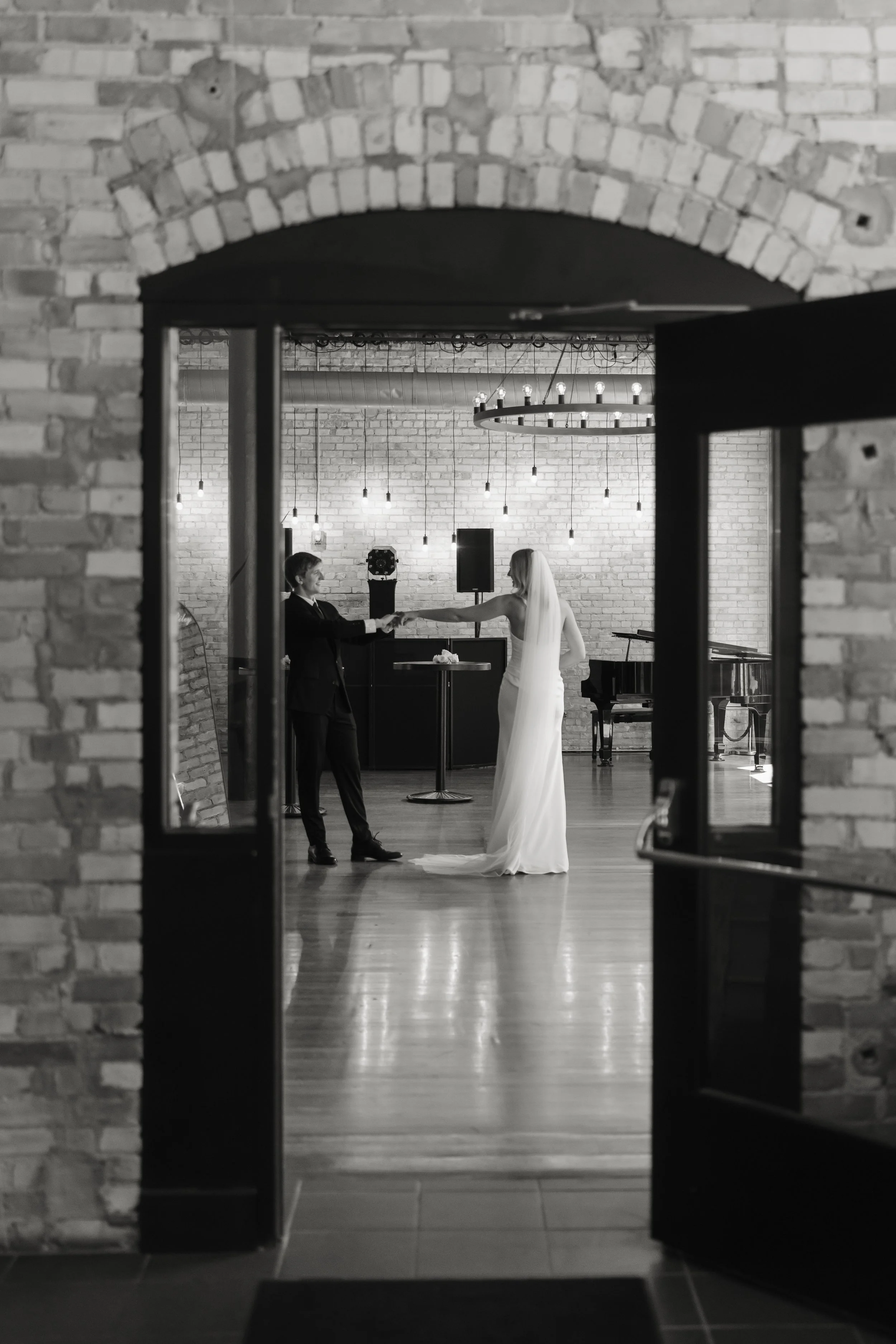 A black and white photo of a wedding ceremony viewed through an open door with a brass archway. The bride and groom are holding hands, facing each other, in a room with a brick wall and hanging lights.