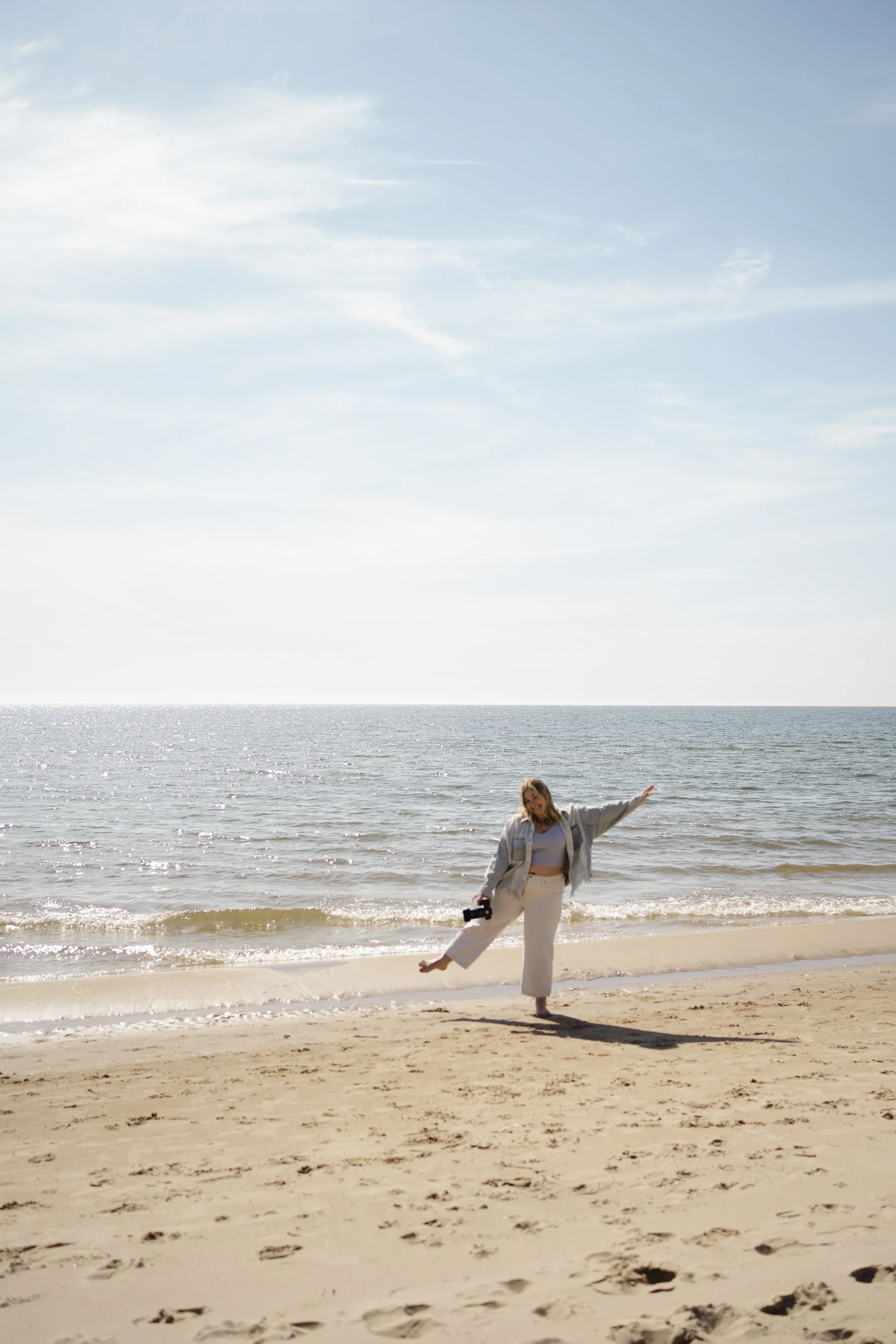 A woman standing on a sandy beach with the ocean behind her, raising one leg and holding a camera in her right hand, smiling at the camera, wearing a light-colored jacket and pants, with a clear sky overhead.