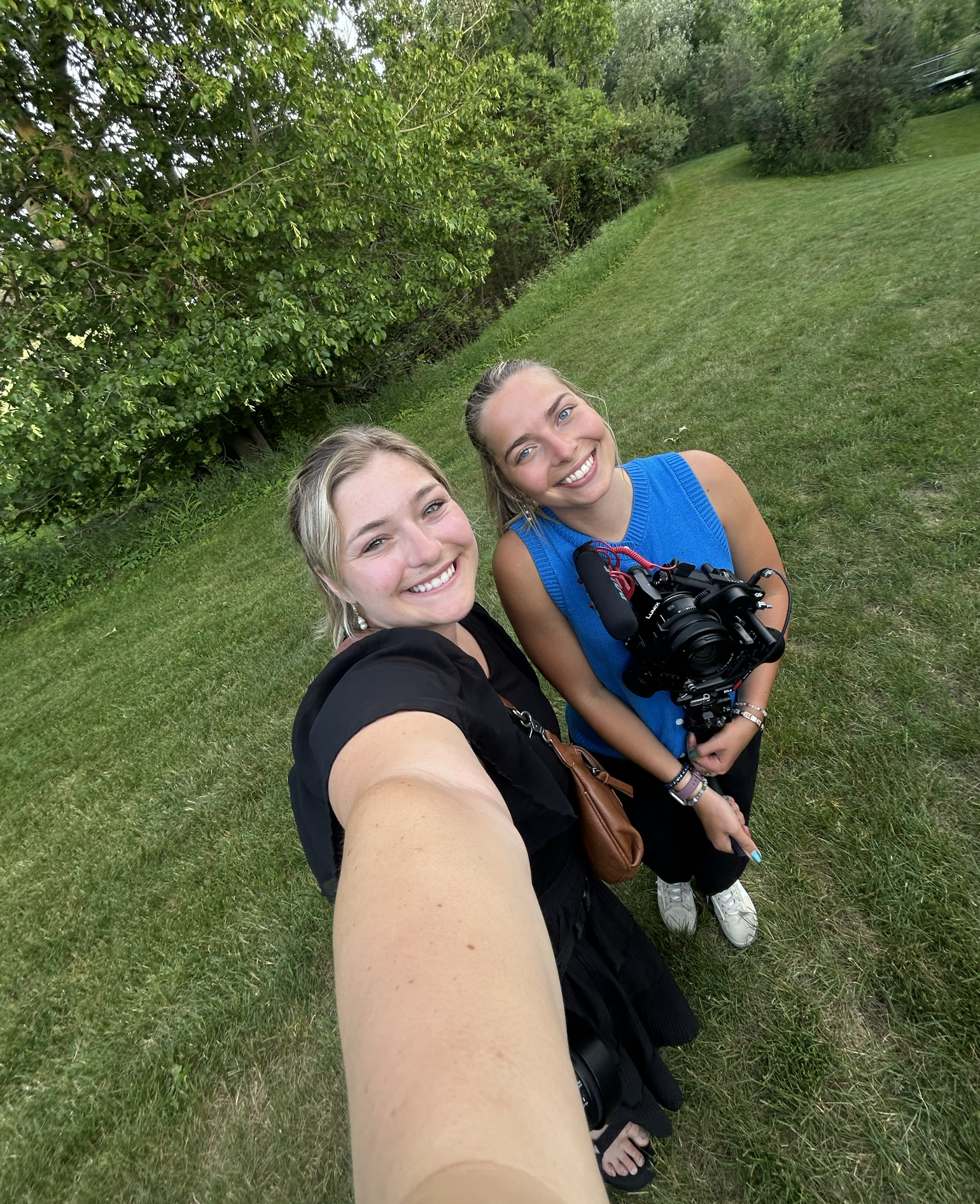 Two women taking a selfie outdoors on a grassy area with trees in the background. They are smiling and looking at the camera, with one woman holding a camera in her hand. One has dark blonde hair and is wearing a black top, and the other has brown ha