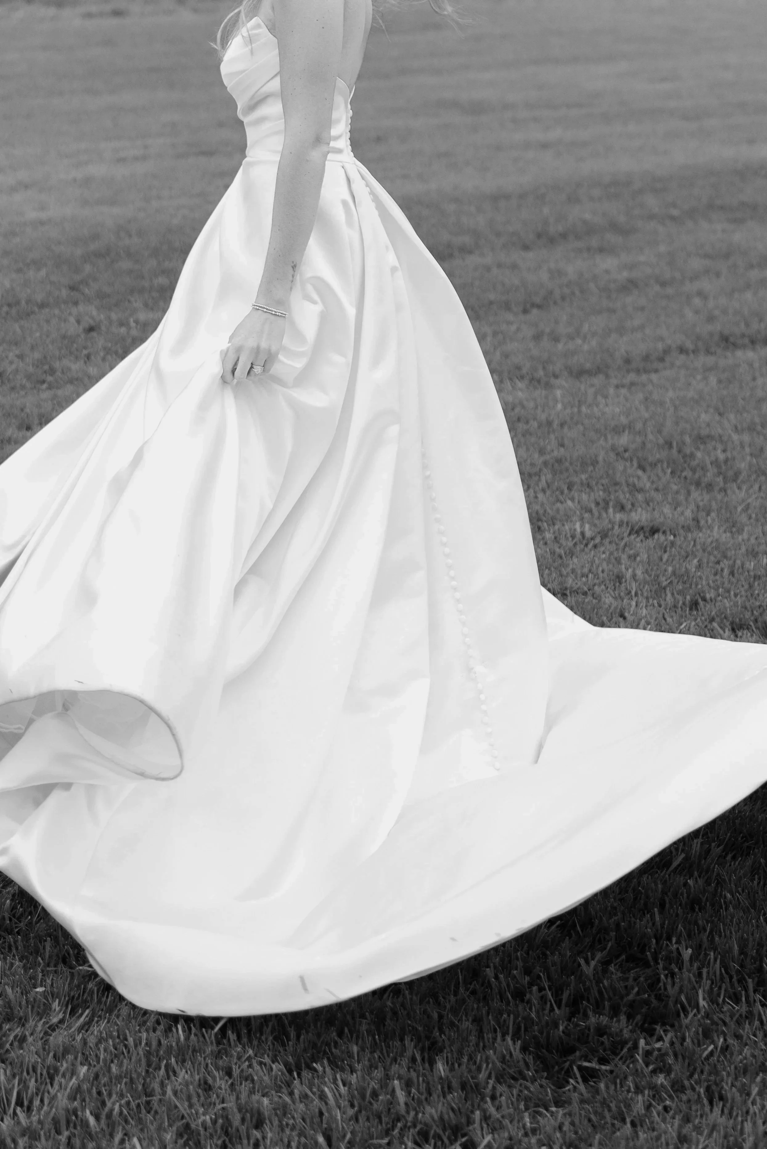 Black and white photo of a woman in a wedding dress standing on grass, with folds and pearls in the skirt visible.