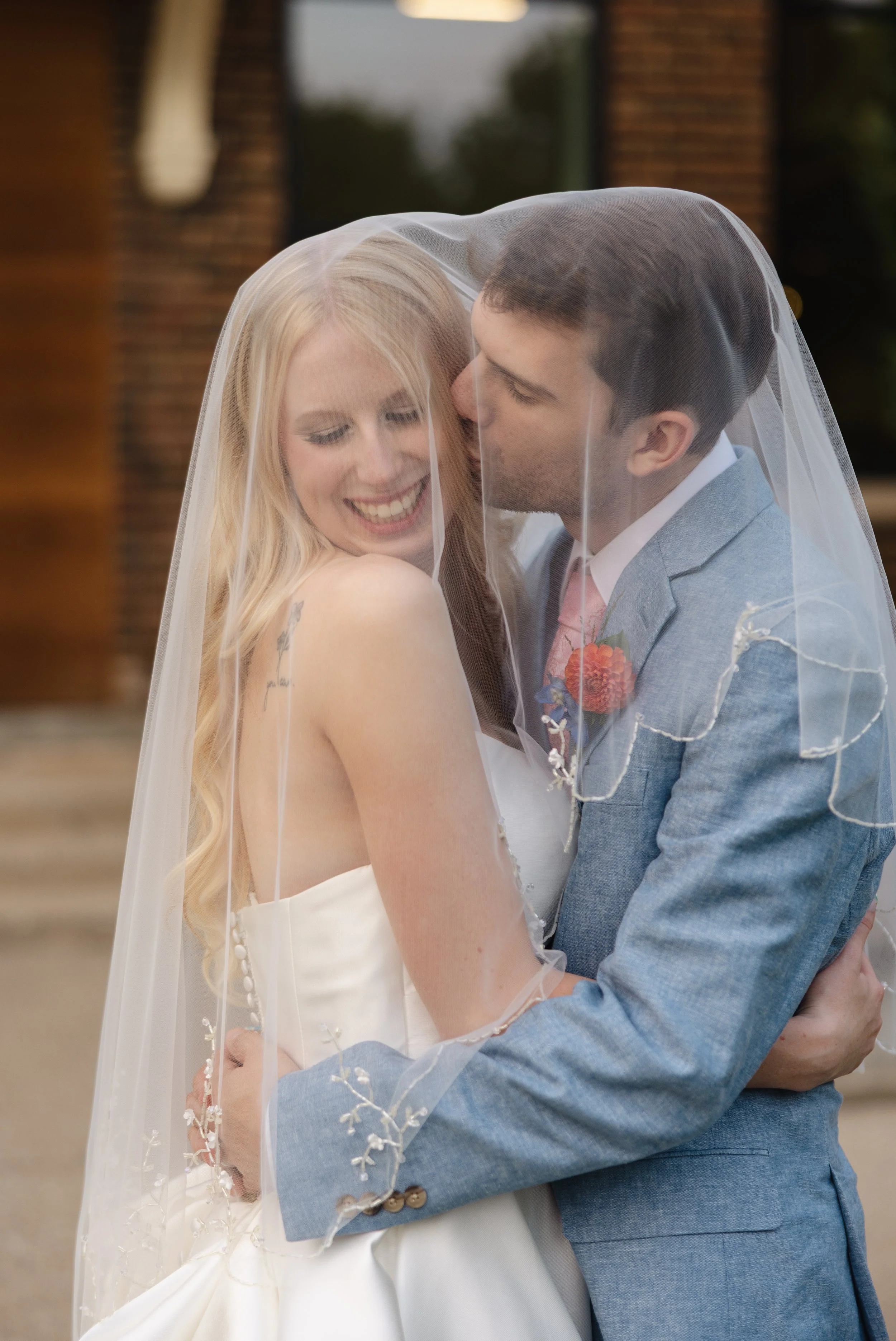 A smiling bride and groom sharing a moment, with the groom kissing the bride's cheek. The bride is wearing a white wedding dress and veil, while the groom is dressed in a light blue suit with a boutonnière. They are outdoors near a brick building.