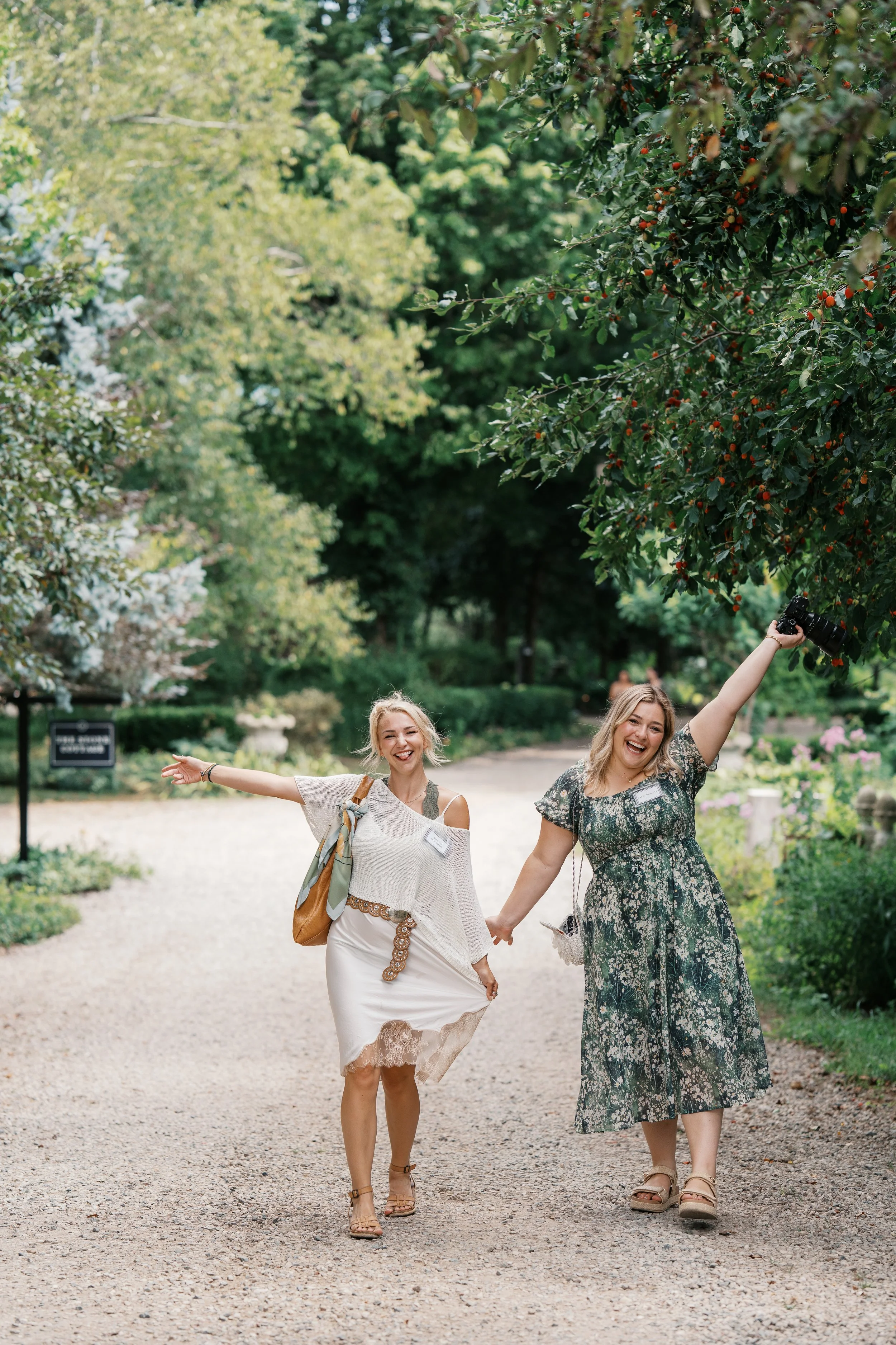 Two women holding hands and smiling, walking on a gravel path in a park with lush, green trees and colorful bushes around them. One woman is holding a camera in the air. They are dressed in casual summer clothing.