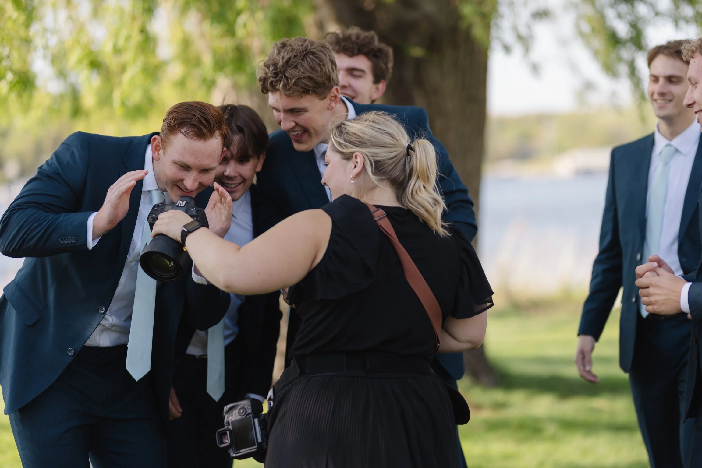 A group of six young adults, dressed in suits, gathered outdoors under a tree, looking at a camera and smiling, with a woman in the foreground and five men around her.
