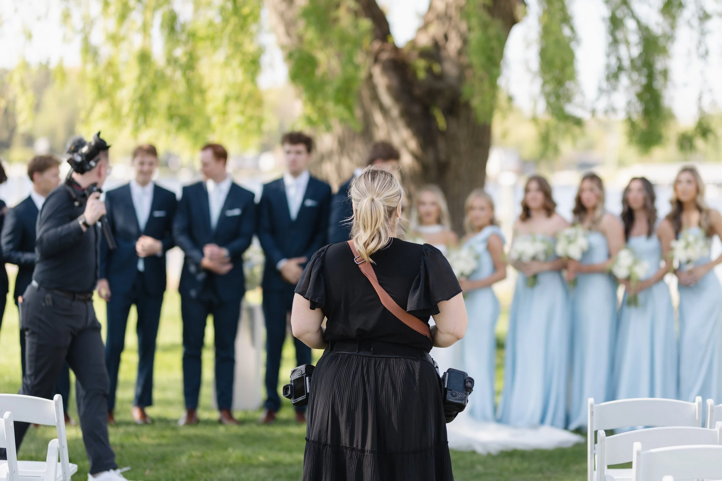 A woman is facing a group of people dressed in formal attire outdoors, with a large tree and green foliage in the background. A man is holding a camera.