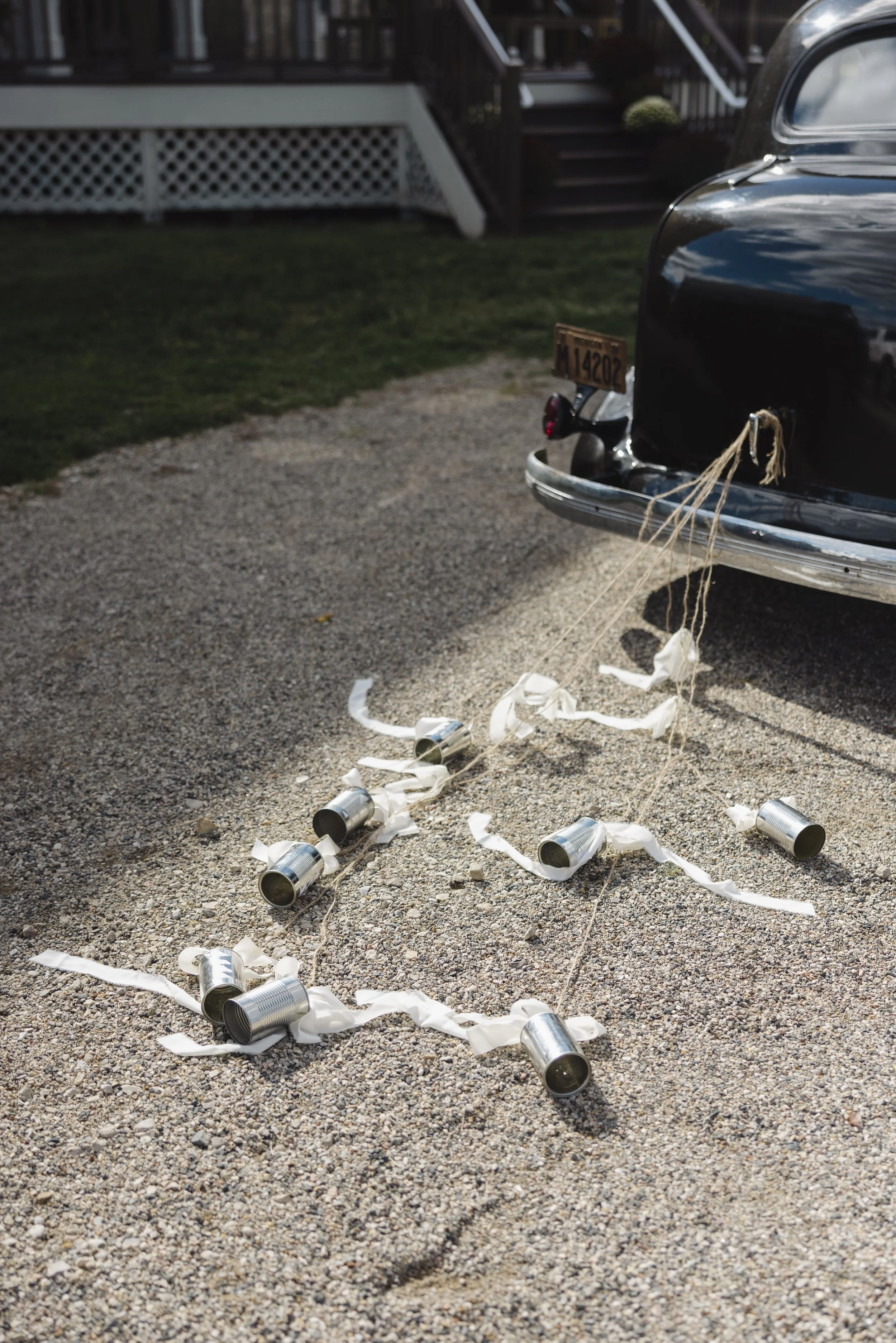 Disorderly scattered tin cans and white paper streamers on gravel driveway in front of a black vintage car with a New York license plate.