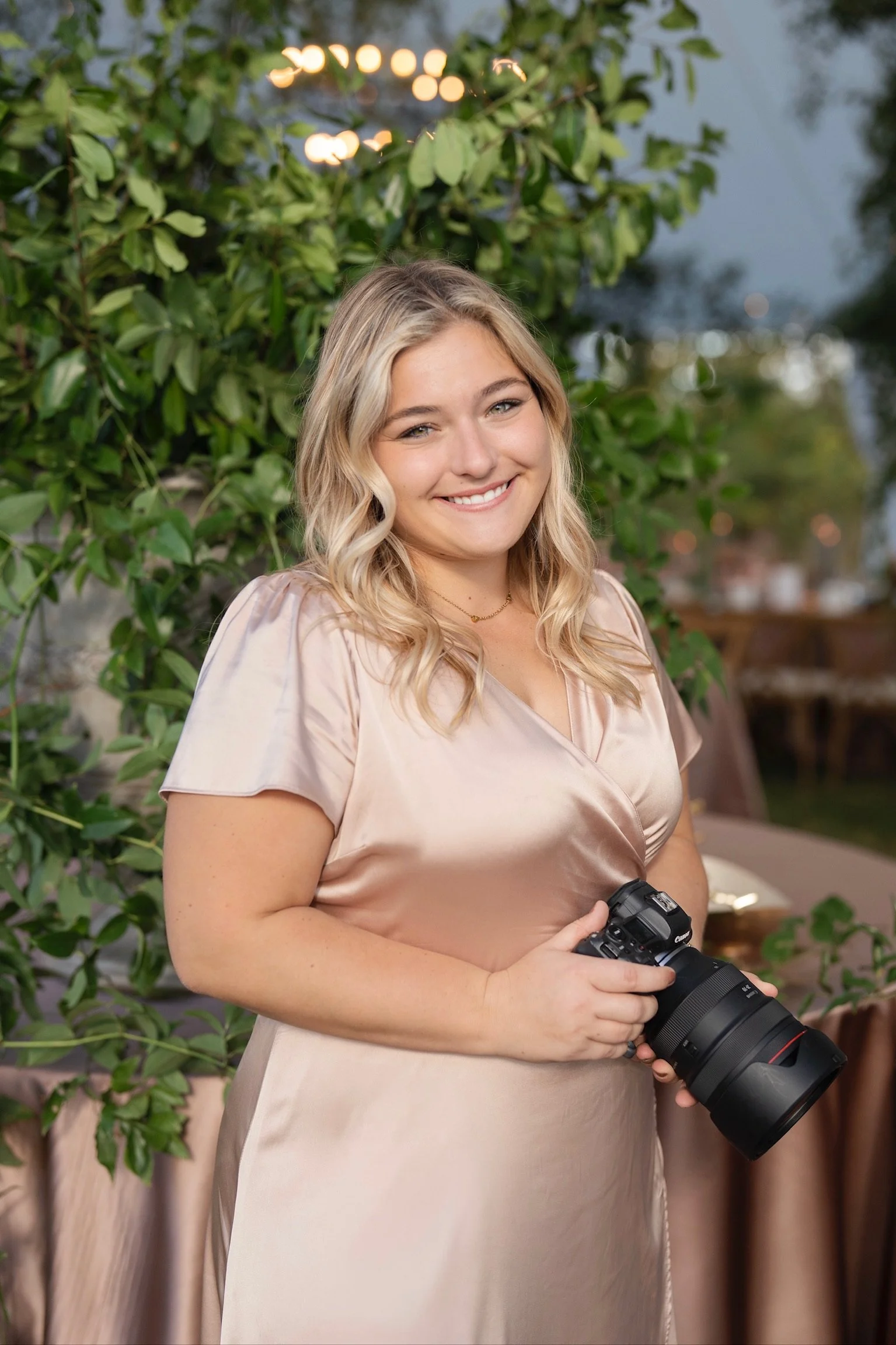 A smiling young woman with wavy blonde hair wearing a silky beige dress, holding a professional camera, standing outdoors in front of green foliage and blurred lights.