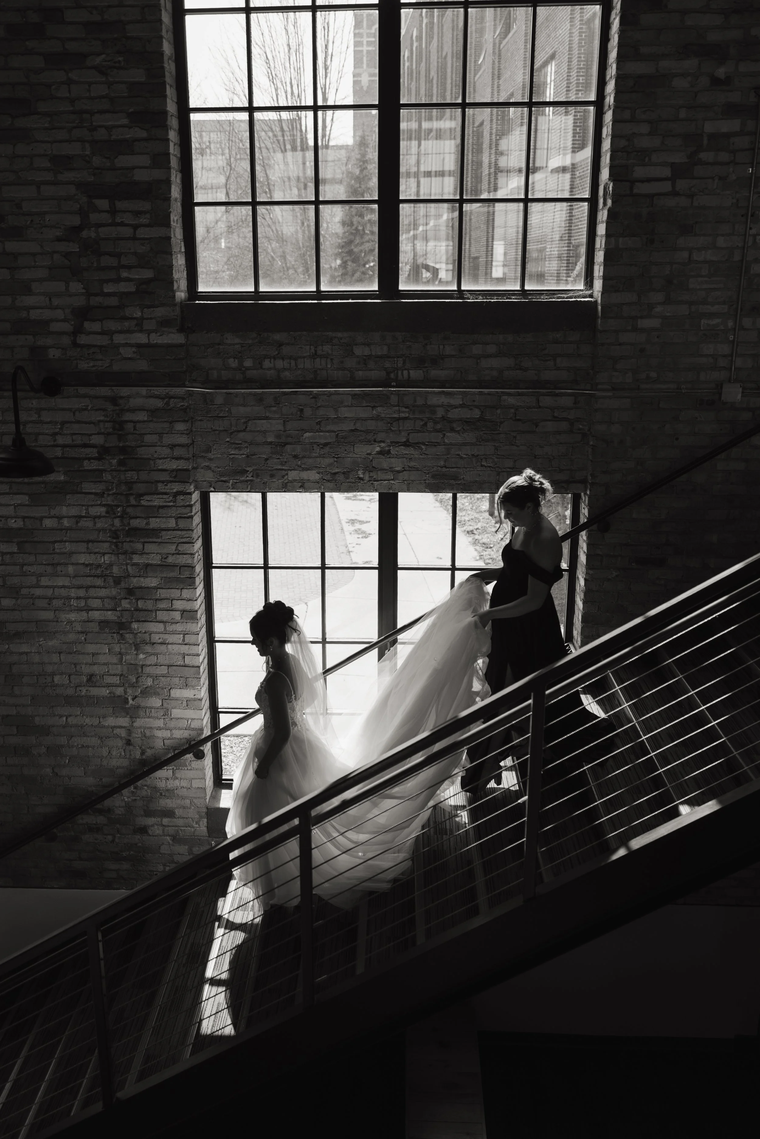 Black and white photo of a woman in a wedding dress walking down the stairs, with another woman helping her with the train, in a space with large windows and exposed brick walls.