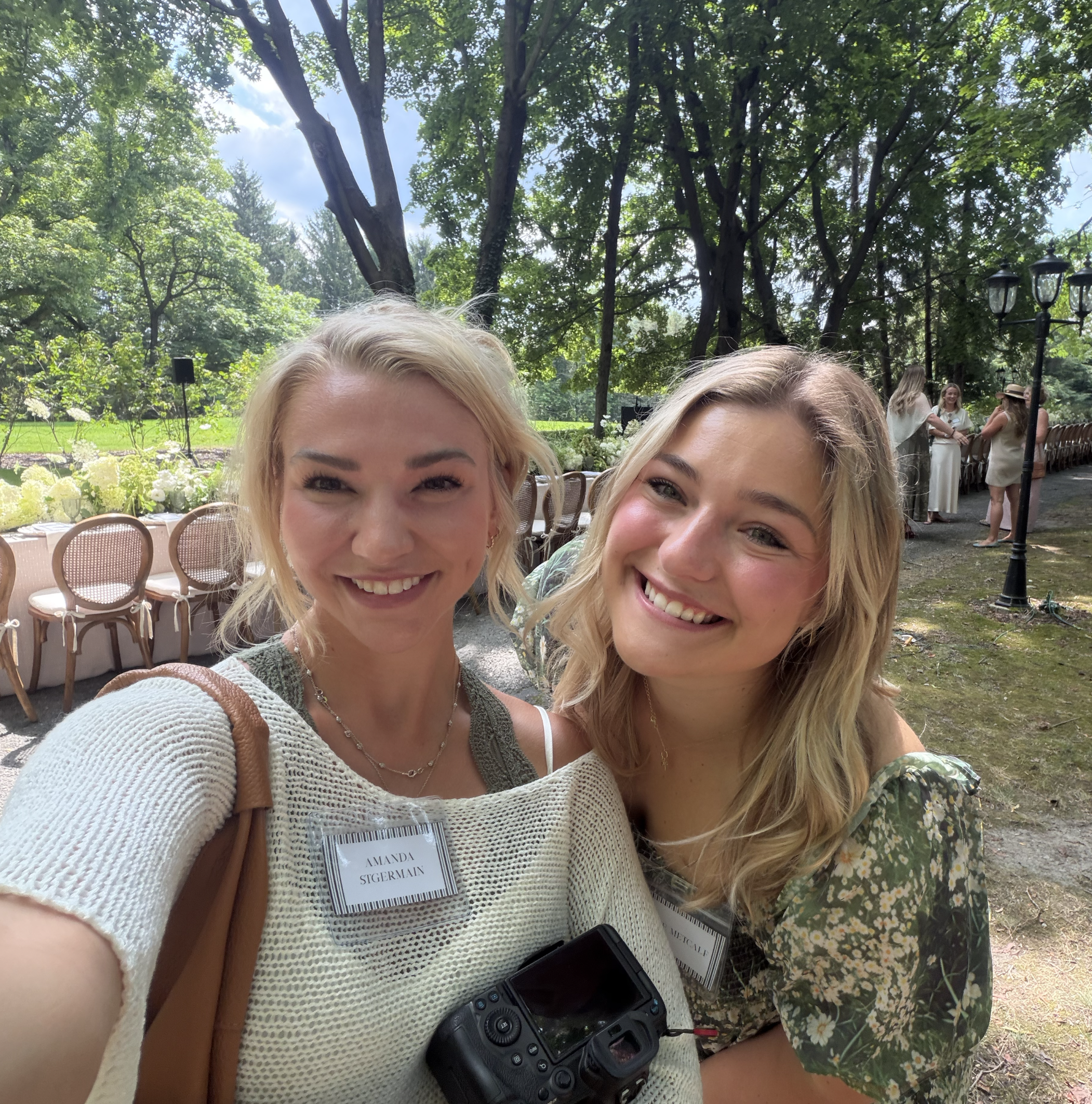 Two smiling women with blonde hair taking a selfie outdoors in a lush green park. One woman has a name tag that reads 'Amanda Stigermain', and a camera or recording device hangs around her neck. In the background, there are trees, a lamppost, and oth